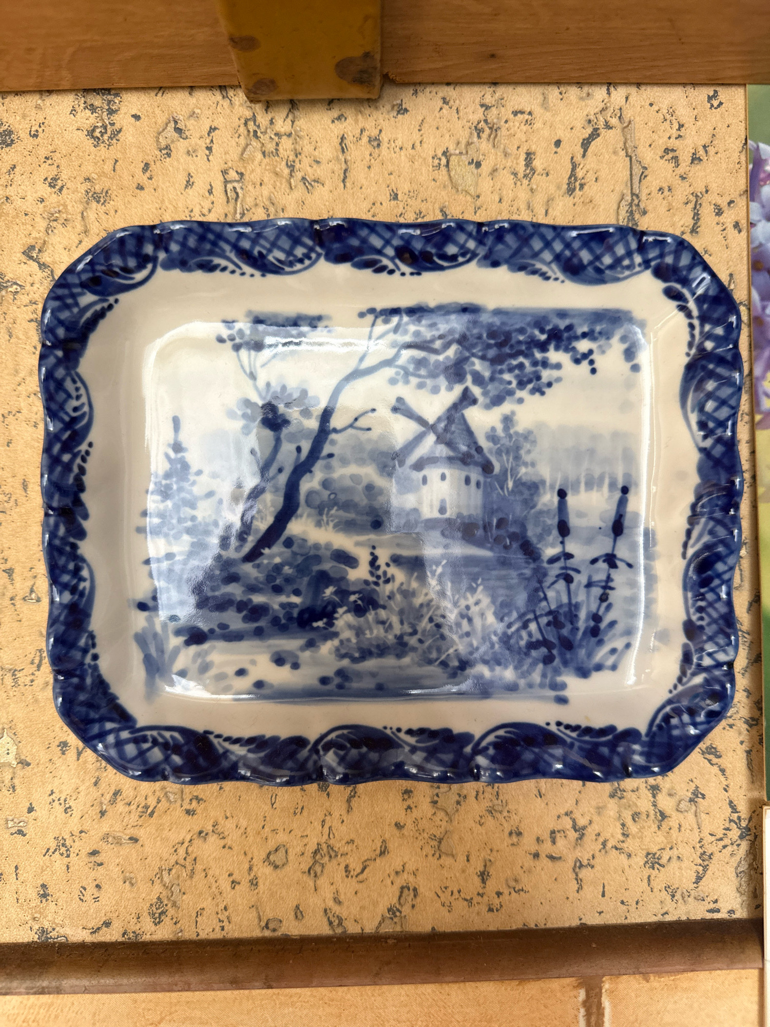 An overhead shot shows a rectangular, white ceramic dish with a decorative blue rim. The dish is placed on a textured, golden-brown surface, possibly cork or a similar material. The rim of the dish features a repeating pattern of blue swirls and dots. The interior of the dish is adorned with a blue and white landscape painting. The painting depicts a windmill with a thatched roof situated in a rural setting with trees, bushes, and reeds. The painting style is suggestive of traditional blue and white pottery. Part of a wooden surface and a yellow object are visible at the top of the frame, and a sliver of a colorful floral image is present on the right side. There are no people or animals in the image. The specific location in Rudamina, Lithuania is not discernible from the image.
