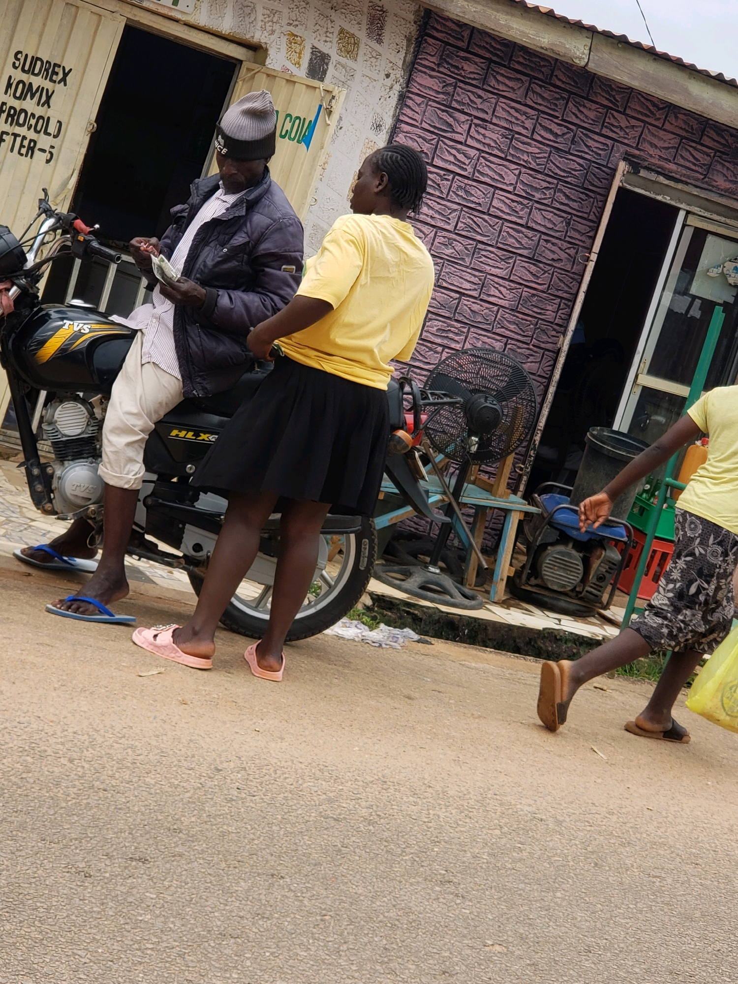 The image shows a street scene in Narayi, Nigeria. Two men and two women are present. One man, wearing a dark jacket and a grey beanie, is seated on a black motorcycle and appears to be counting money. He is wearing light-colored trousers. Beside him, a woman in a yellow t-shirt and a black skirt stands. To the right of the motorcycle, another woman, also in a yellow t-shirt, is walking away from the viewer, carrying a yellow plastic bag. She is wearing patterned shorts. The background includes a building with a corrugated iron roof and walls with a brick pattern. A large black oscillating fan is visible behind the motorcycle. A blue and yellow generator is also situated near the building. The ground is paved with asphalt. The scene suggests a commercial area, possibly a roadside stall or shop.