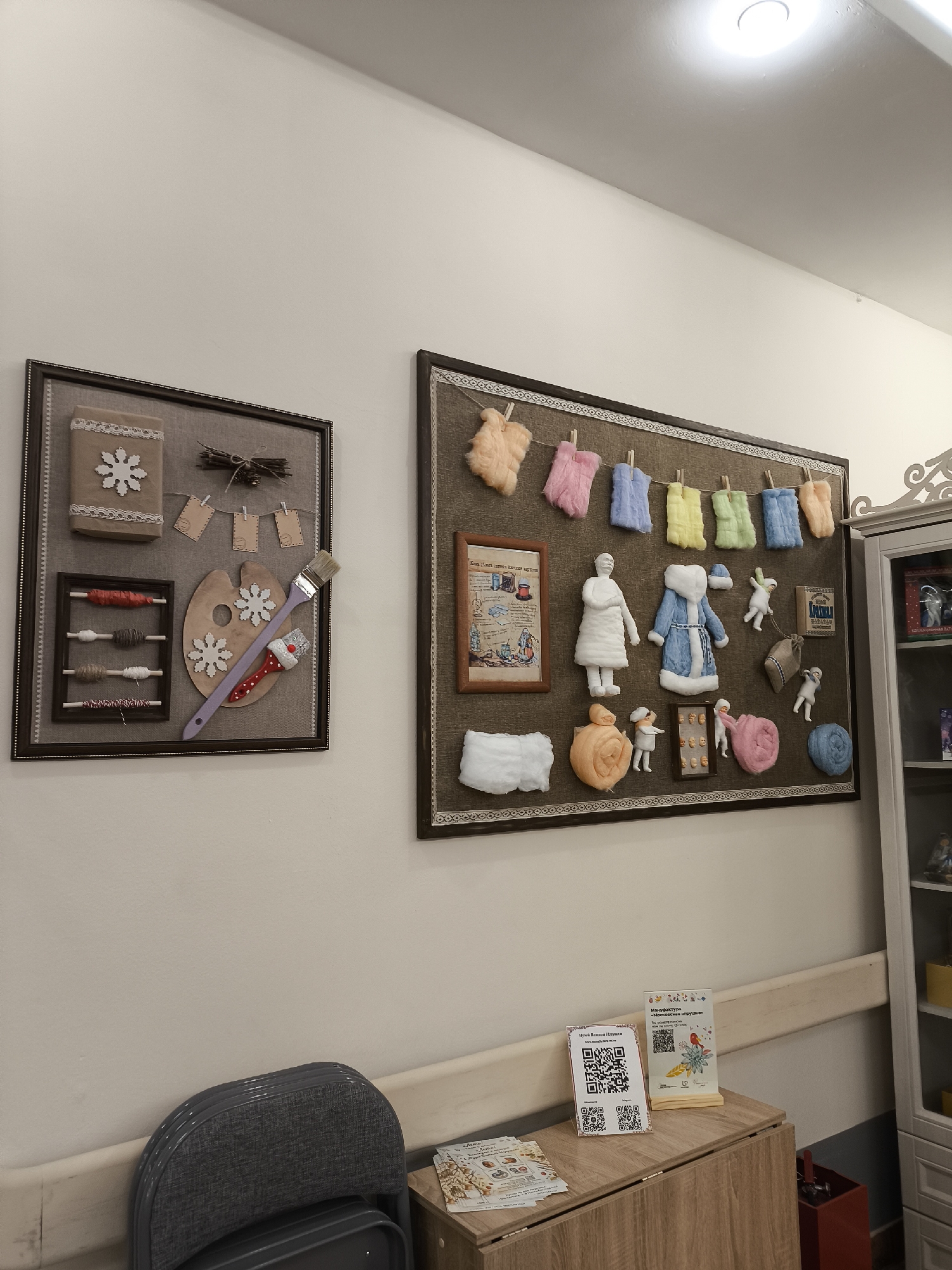 The image shows a wall in what appears to be an indoor space, likely a craft shop or display area, somewhere in Moscow, Russia, based on the text on the displayed items.
Two framed artworks hang on the wall. The one on the left features craft items, including a fabric-covered block decorated with a snowflake, a small twig bundle, miniature clothespins hanging tiny cards, an artist's palette with snowflake decorations, an abacus with colored yarn, and a paintbrush. The artwork on the right contains a similar craft display, including small fabric or felt representations of clothing items hanging on a string, figures, and a framed document or image with Cyrillic text.
Below the artworks, there's a storage unit with a light wood-grain finish. On top of the unit are two small informational displays with QR codes and Cyrillic text. A gray, folding chair is positioned beside the storage unit.
To the right of the unit is a tall, white cabinet, parts of which are visible, displaying some items on its shelves.
The walls are painted a neutral, light color. The lighting is provided by a ceiling-mounted light fixture. The scene gives the impression of a creative and potentially educational or commercial space.