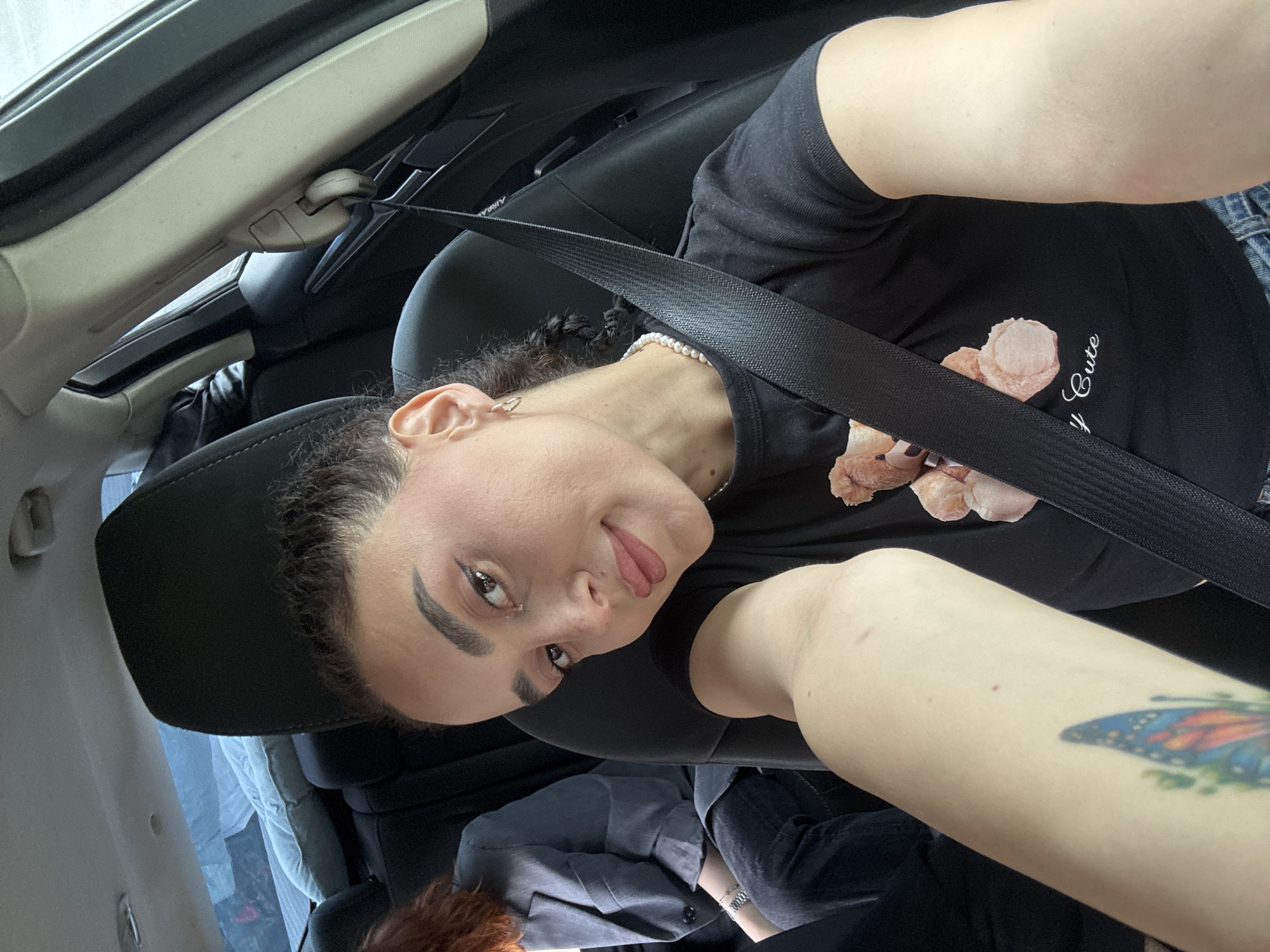 A person with fair skin, dark hair, and visible makeup is seated in the driver's seat of a car. They are wearing a black t-shirt with a light-colored floral design and a pearl necklace. A seatbelt is fastened across their body. They are holding the camera at arm's length, taking a selfie. A butterfly tattoo is visible on their arm. The car's interior is visible, including the seat, dashboard, and roof. An individual is visible in the bottom corner.
The location is not discernible from the image.