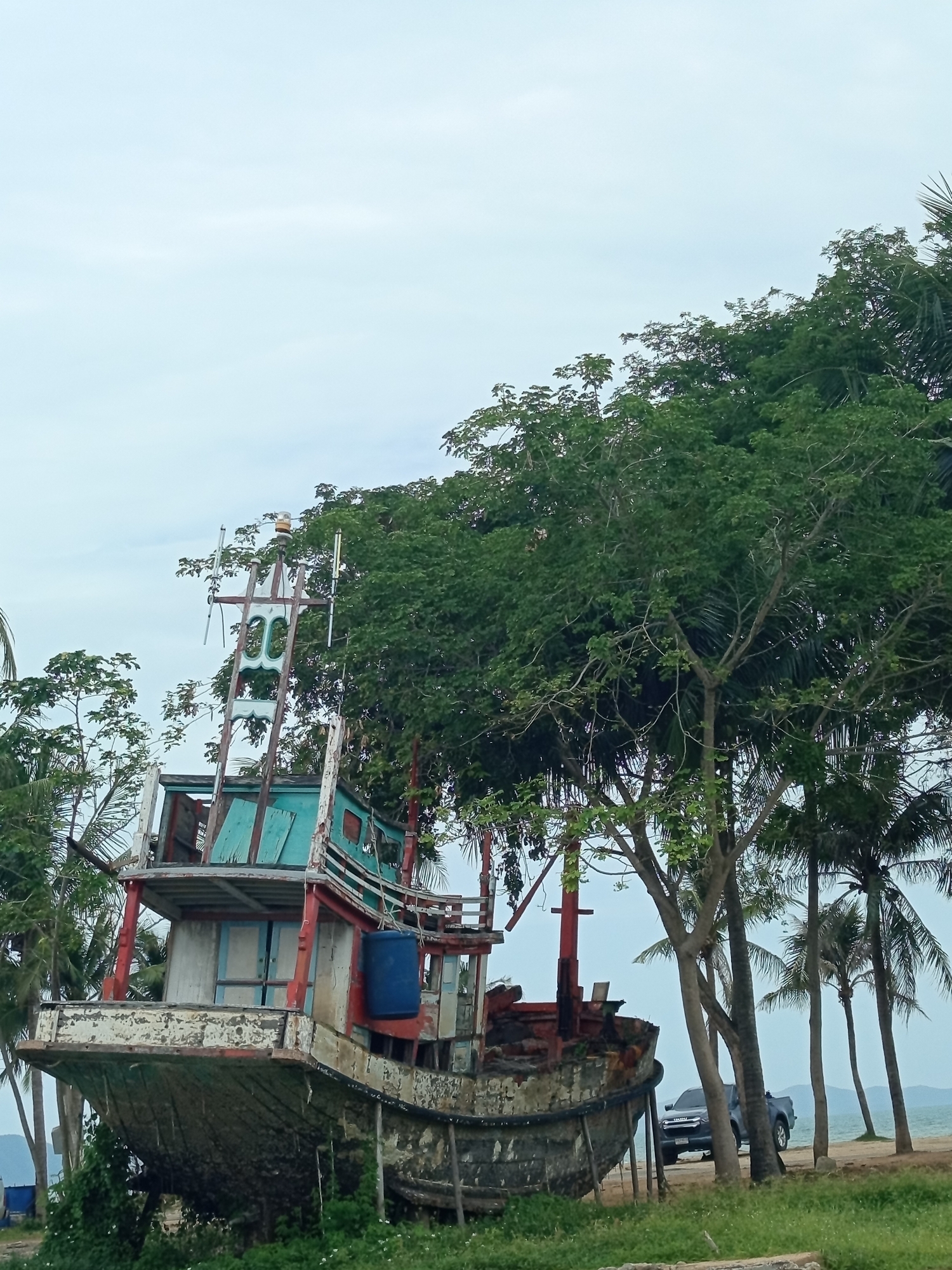 This is a full-frame, eye-level, outdoor, medium shot of a beached and decaying fishing boat in Ban Na Chom Thian, Thailand. The boat, which is mostly gray with peeling white, red, and turquoise paint, is partially submerged in thick green foliage and tilted at an angle. It has multiple levels, a blue barrel attached to its side, and a tall mast with lights and what appears to be antennae. In the background, palm trees and other lush green trees are visible. A dark colored pickup truck is parked on a sandy area to the right of the boat, near the shoreline. The sky is overcast and light blue. The overall scene depicts a forgotten vessel amidst abundant vegetation near the sea.