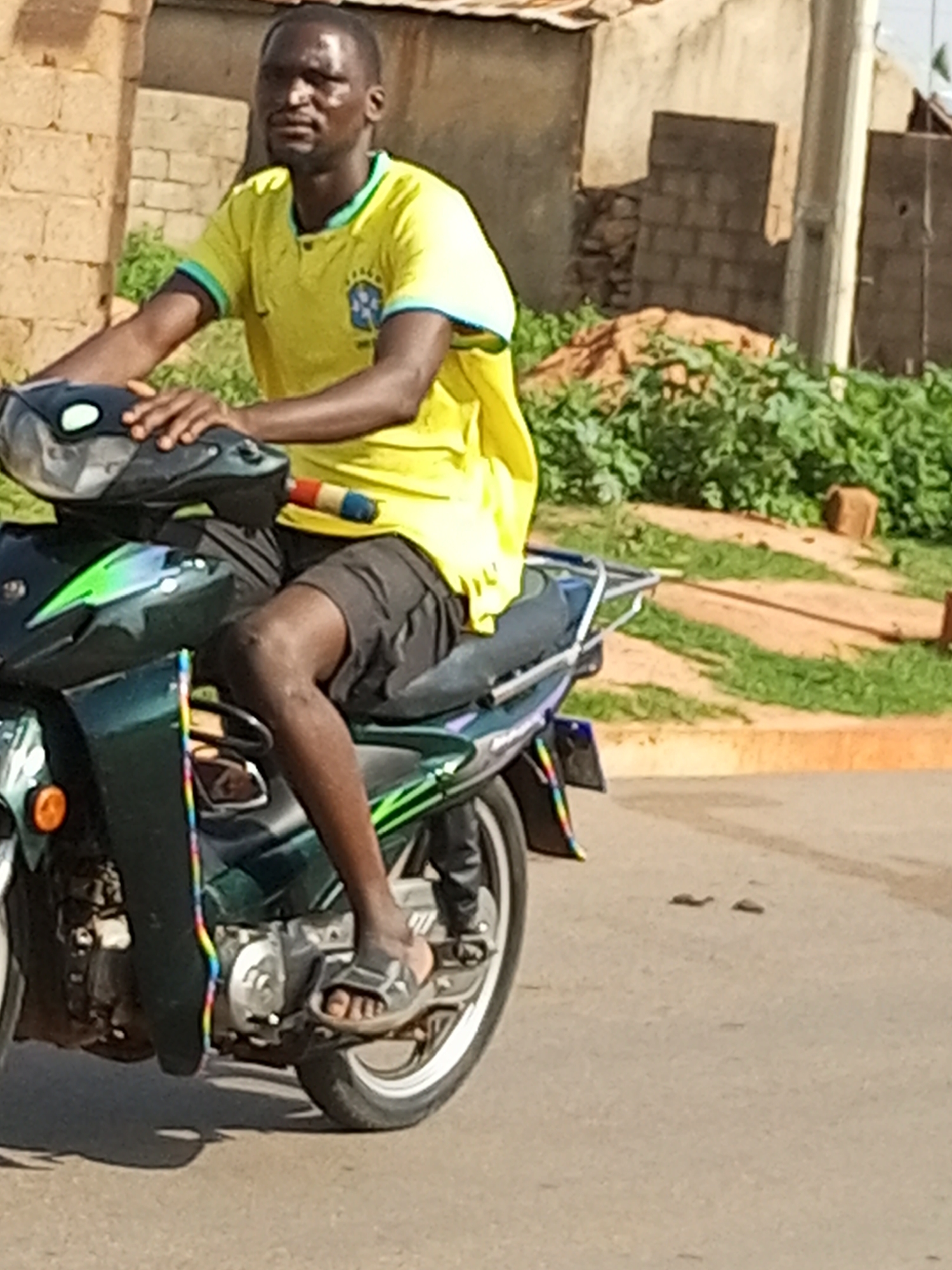 A man, appearing to be of African descent with dark skin and a shaved head, is seated on a dark green motorcycle. He is wearing a bright yellow short-sleeved t-shirt with a logo on the left chest, and dark shorts. His right hand rests on the handlebars, and his left arm is bent, resting on the front of the motorcycle. His legs are straddled over the motorcycle, with his right foot wearing a dark sandal resting on the footrest. The motorcycle has a dark front panel with green accents and a light-colored front fender. A colorful ribbon-like decoration is visible on the side of the motorcycle. The background shows a rough, tan-colored wall with some brickwork, and beyond that, a densely vegetated area with green foliage and scattered brown objects that appear to be bricks. The ground is paved and appears to be a road. The scene suggests an outdoor environment in a built-up area, possibly in the city of Gombe, Nigeria.
