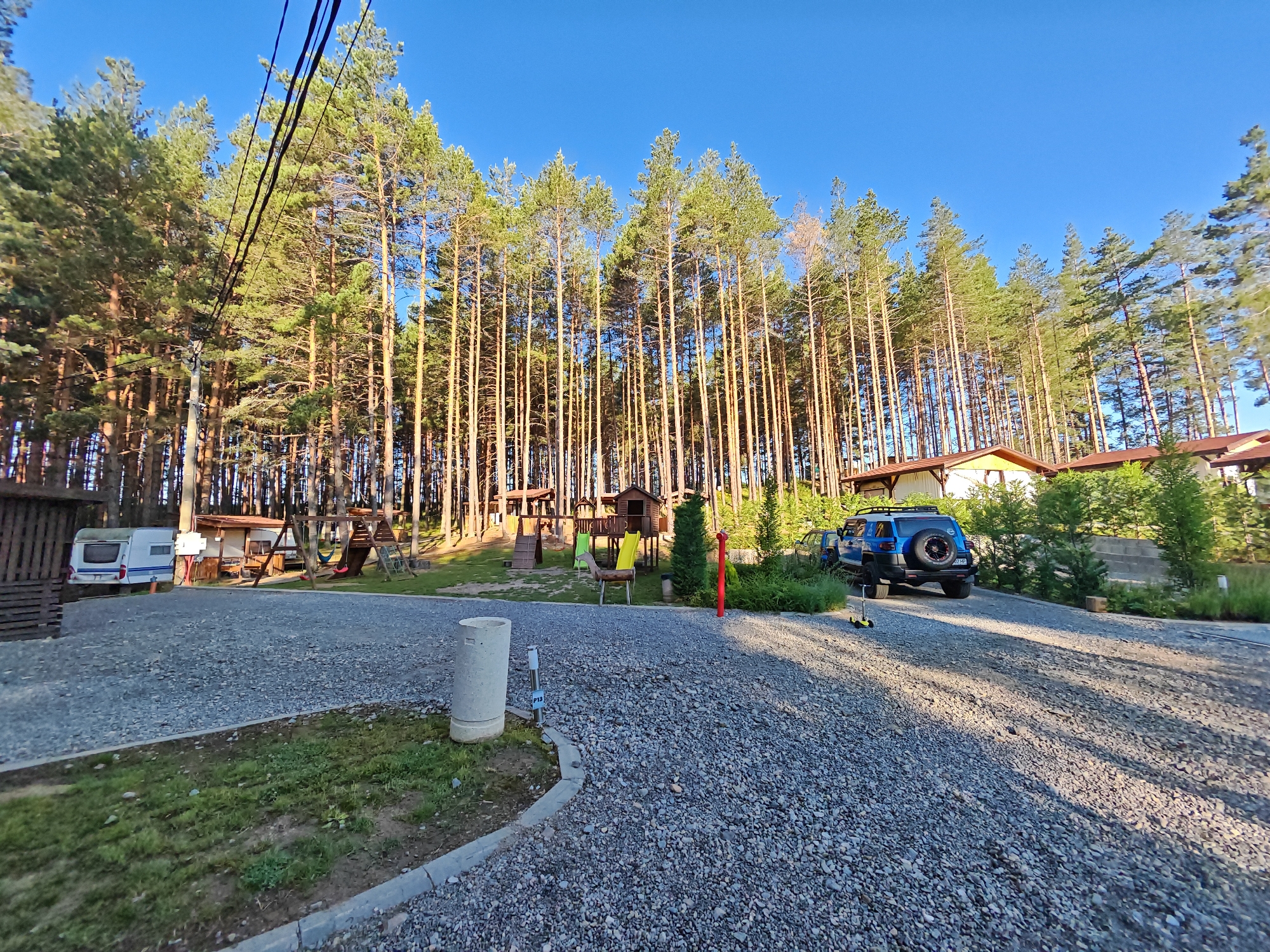The media depicts an outdoor scene on a sunny day, likely in the late afternoon given the long shadows cast by the trees. The setting is a wooded area with a clear blue sky. In the foreground, a gravel driveway curves through the scene. To the left, a white caravan is parked. Further back, a children's playground with a wooden fort, slide, and swings is visible amongst the trees. Several tall pine trees dominate the background, creating a dense forest. A blue SUV is parked on the gravel area to the right of the playground, and a yellow scooter is nearby. Behind the playground and SUV, there are buildings with brown roofs, suggesting a residential or camping area. The overall atmosphere is peaceful and recreational, hinting at a holiday or camping destination. The ground is a mix of gravel and patches of grass. There is a concrete cylindrical object with a sign attached in the foreground, which appears to be a trash receptacle or marker, with the text "P13" visible on the sign. No people are visible in the image. The location is contextually Alino, Bulgaria.
