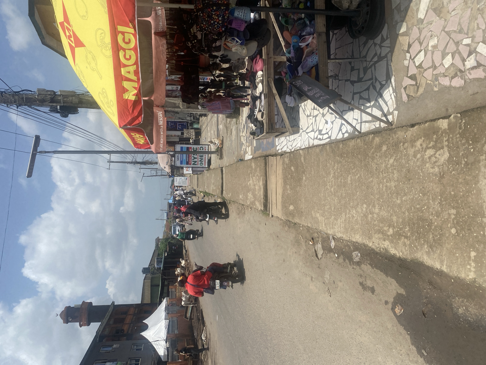 The image depicts a bustling street scene in Lagos, Nigeria. The view is captured from street level, looking down a road lined with shops. The shops feature vibrant awnings and displays, including one with a "MAGGI" advertisement and another with a shoe display and a sign indicating a sales girl is needed. A mosque tower with a crescent moon adorns a building on the left side of the image. Motorbikes are seen traveling down the street, some carrying passengers. Utility poles with power lines are visible against a cloudy sky. The pavement appears worn, and the overall atmosphere is one of an active commercial area.