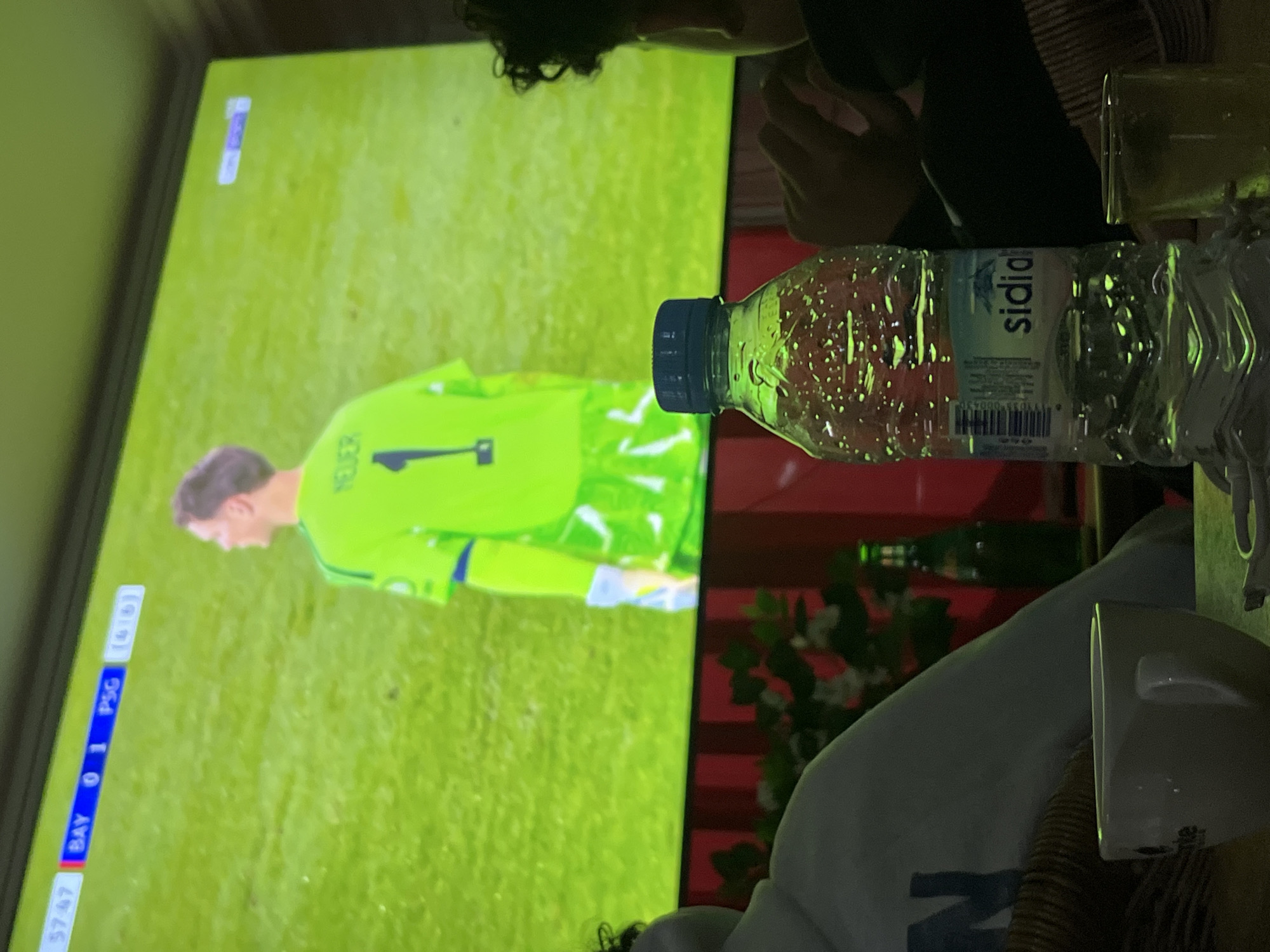 A person is watching a soccer game on television. The screen shows the Bayern Munich vs. Paris Saint-Germain game with the Bayern goalkeeper, Neuer, on the screen. A bottle of Sidi Ali water is in the foreground. The television screen shows the location of Ouaouizert, Morocco.