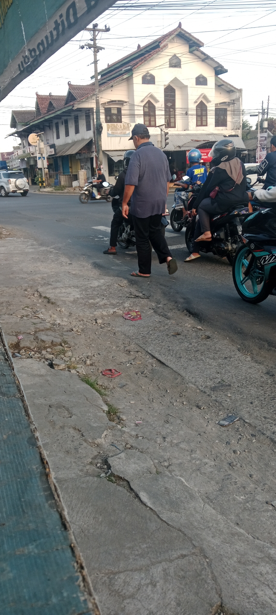 In Maguwoharjo, Indonesia, a street scene unfolds with several individuals on motorcycles and one person walking. The person walking, a man wearing a blue t-shirt and black pants, is captured in mid-stride, his flip-flops visible. He is walking on a cracked concrete surface adjacent to a paved road where several motorcycles are present. One motorcycle, turquoise and black, is prominently in the foreground, its wheel turned. Another motorcycle, ridden by a person in a black hijab and grey top, is stopped. Further back, a motorcycle with a blue helmet and a rider in a blue shirt is also stopped. A white SUV is seen turning at a junction in the background. The buildings in the background are a mix of traditional architecture with red-tiled roofs and more modern facades, some with signs indicating businesses. The sky is bright, suggesting daylight. The concrete ground is littered with debris, including a red and yellow object and a single red flip-flop.