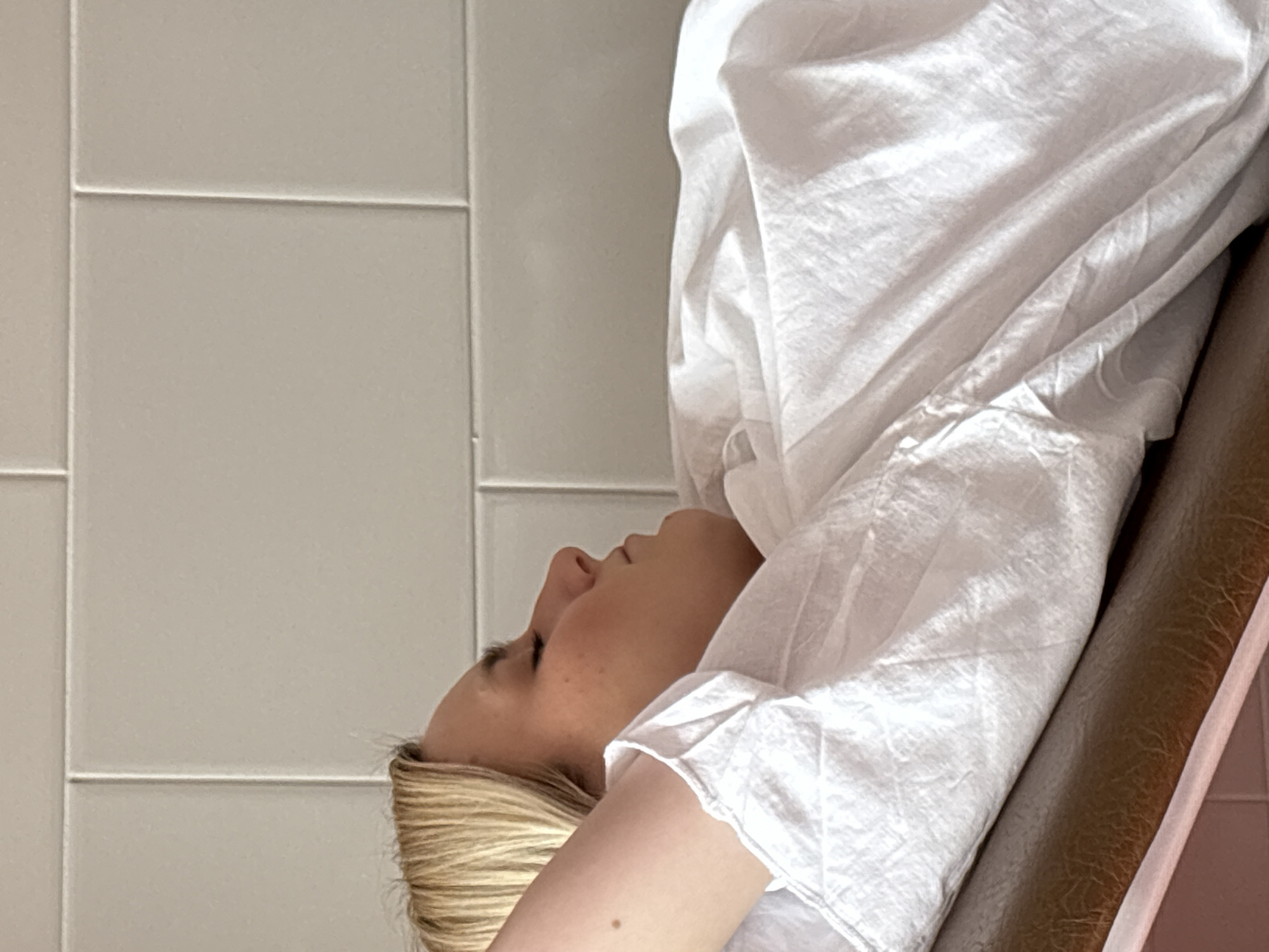 A fair-skinned woman with blonde hair is lying on a brown leather examination table in a room with white tiled walls. She is covered in a white, crinkled paper sheet. The location cannot be determined to be Saint Petersburg, Russia, from the provided image alone.