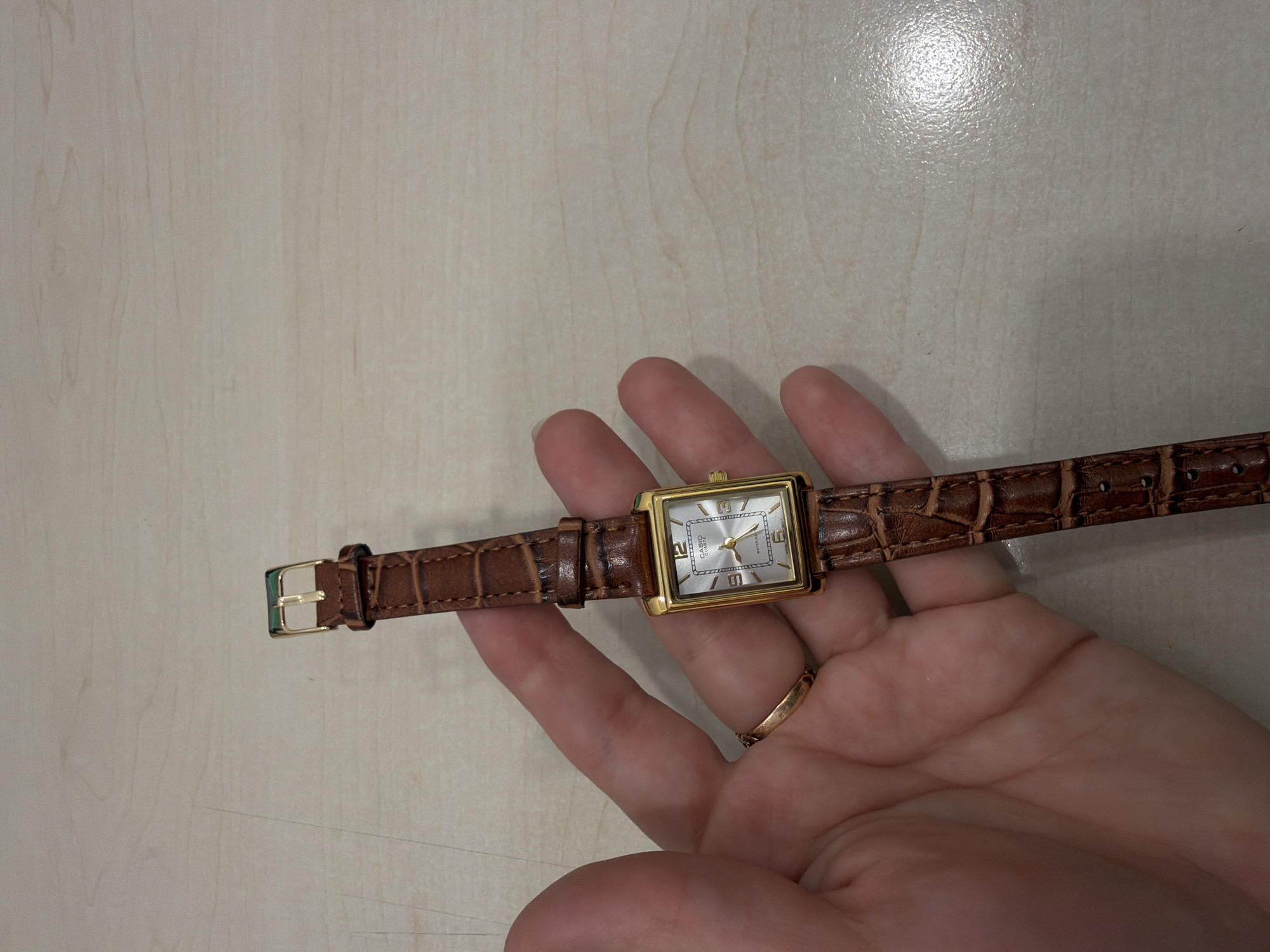 A hand holds a gold-cased Casio watch with a brown leather strap. The watch face is silver with gold numerals. The background is a light, wooden-patterned surface. A gold ring is visible on the hand.