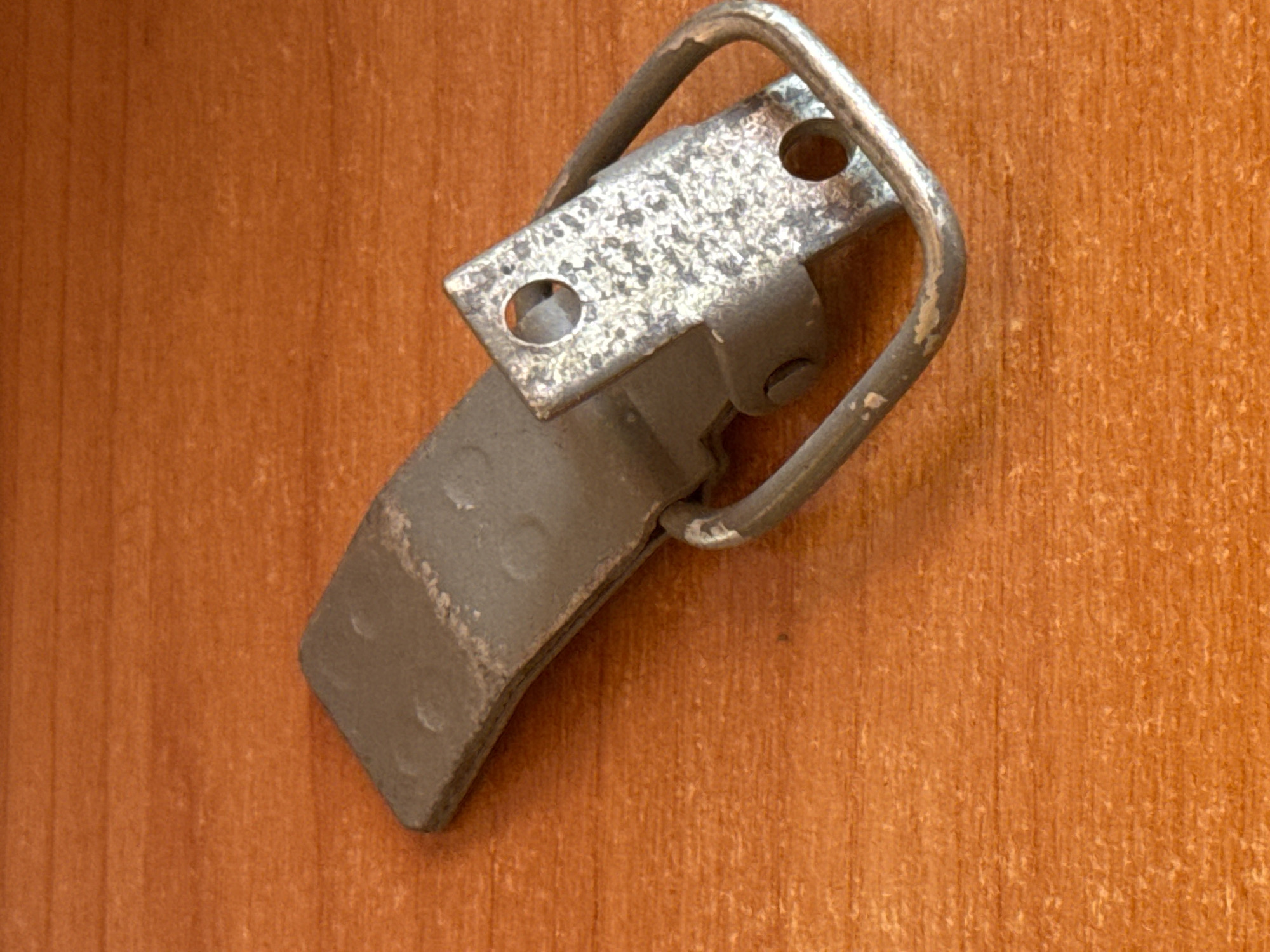 A metal latch, with a loop for a padlock, rests on a textured orange surface that appears to be wood. The latch is silver and shows signs of wear and rust. The wood surface has a prominent grain pattern. The image is an overhead shot, with the latch positioned diagonally across the frame. There are no people or animals present.