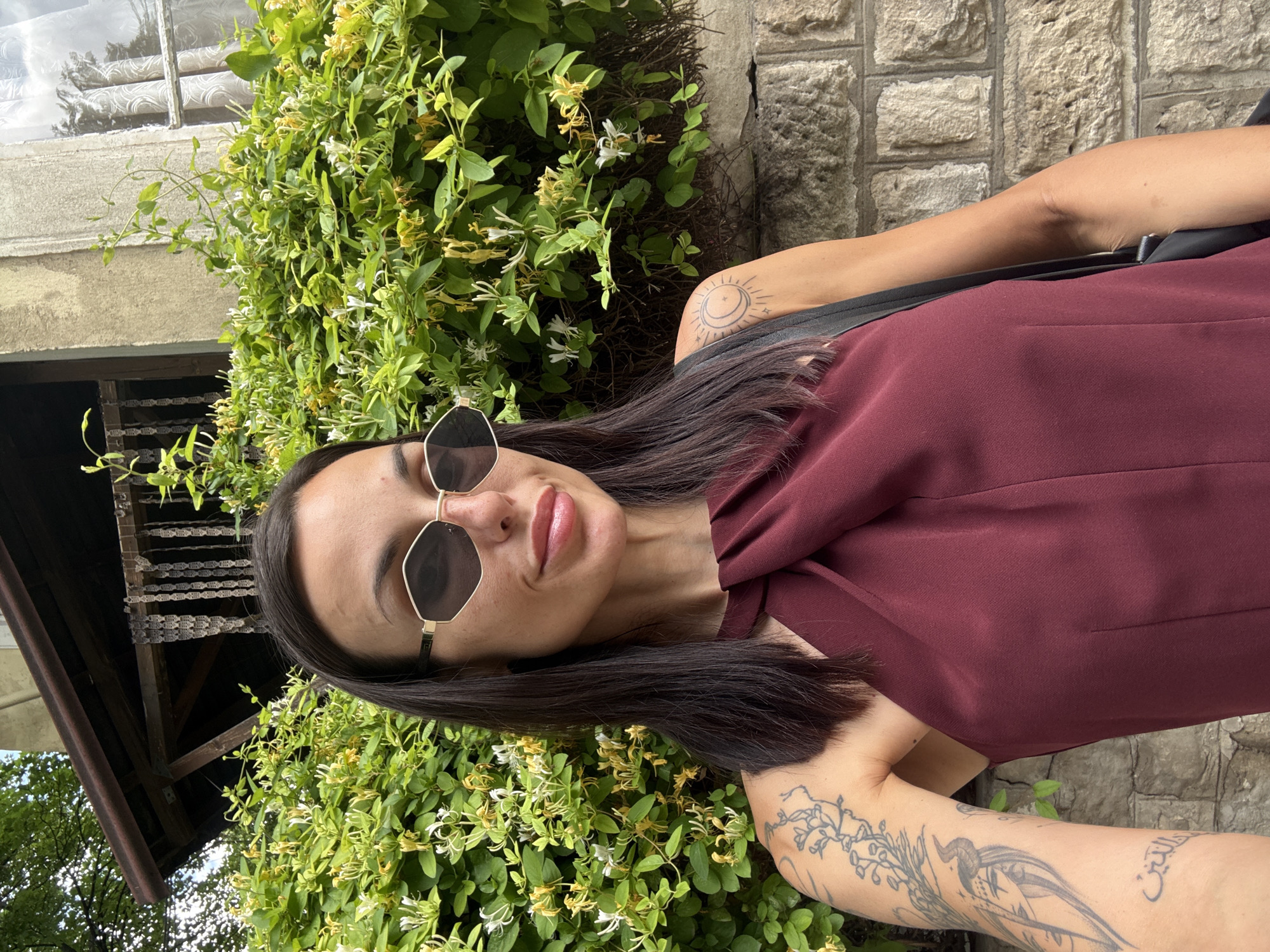 A woman with long dark hair, wearing sunglasses and a maroon top or dress, poses for a photo outdoors in Pleven, Bulgaria. Her head is tilted to the right, and she looks towards the camera with a slight smile. The sunglasses have distinctive octagonal, gold-colored frames.
She has several visible tattoos: a design incorporating a crescent moon, sun, and stars on her right upper arm, and an intricate pattern of foliage, a bird, and text in Arabic script on her left forearm.
The background features lush green foliage with numerous white and yellow flowers, likely honeysuckle, suggesting a natural or garden setting. Behind the foliage, a textured stone wall made of light-colored, rough-hewn blocks is visible. On the far left, elements of a building can be seen, including a window with blinds or shutters and a wooden eave structure, with hints of blue sky and tree branches beyond. The scene is brightly lit, indicating daytime and clear weather.