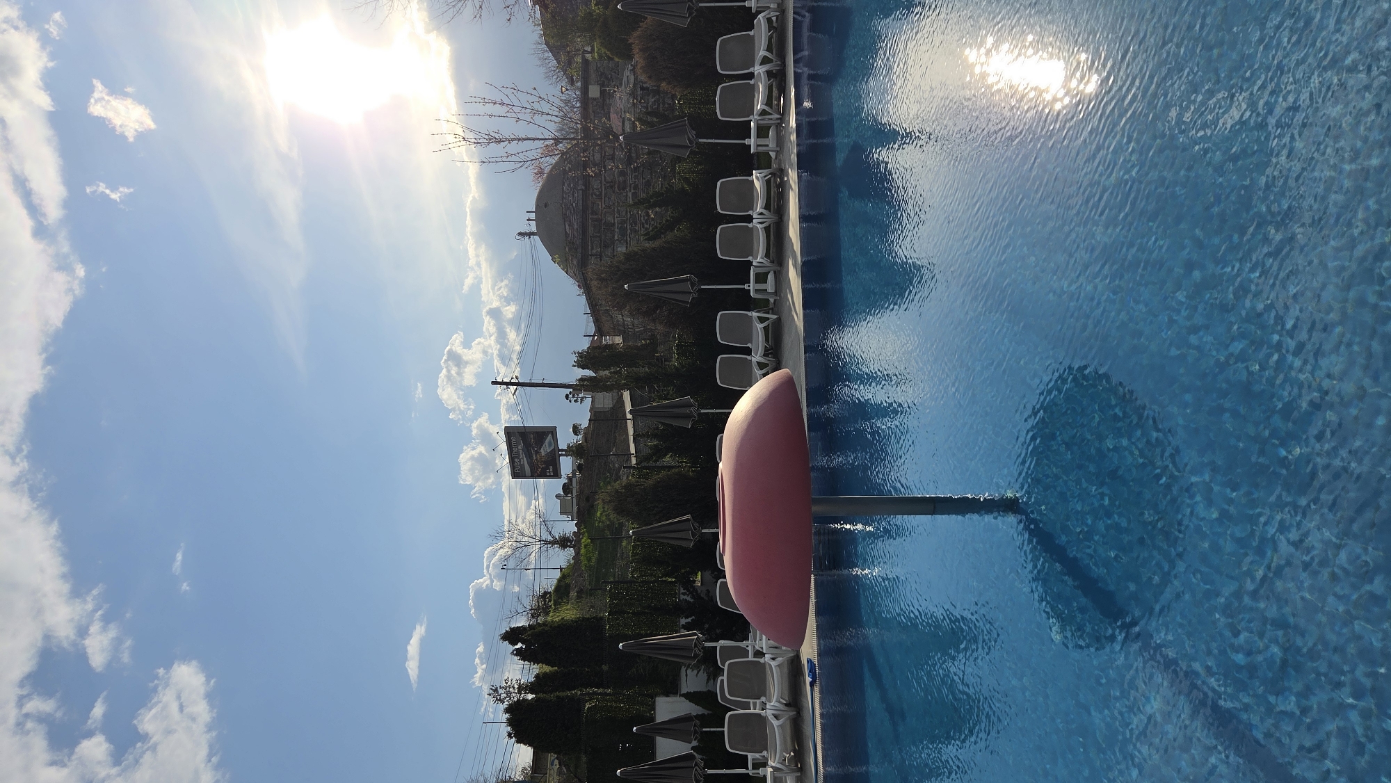 An outdoor scene captures a bright, sunny day at a swimming pool. The image is oriented vertically, with the sky above, the poolside and surrounding landscape to the left, and the vibrant blue swimming pool occupying the right side.
The main subject is a large swimming pool with clear, rippling blue water. The sun reflects brightly off the water's surface, creating dazzling patterns. A distinctive red or pink, smooth, oblong object, possibly a pool slide or a large float, is situated at the edge of the pool. In one of the cropped views, a pole, likely a cleaning tool, is partially submerged in the water.
To the left of the pool, a relaxation area features several white lounge chairs shaded by dark umbrellas, arranged in rows. Beyond this, a strip of green foliage, consisting of various trees and bushes, separates the pool area from the background. In the distance, hills or mountains are visible, dotted with buildings. A prominent feature on one of the hills is a large, grey, domed structure, which could be an old bathhouse or another type of historical or industrial building. Utility poles and wires are also present in the mid-ground. A billboard sign, partially visible, displays the text "Pulse Thermal".
The sky is a clear, bright blue with scattered white, cumulus clouds. The strong sunlight indicates it is daytime, likely a morning or afternoon, given the intensity of the light. No people are present, lending a tranquil and serene atmosphere to the scene.