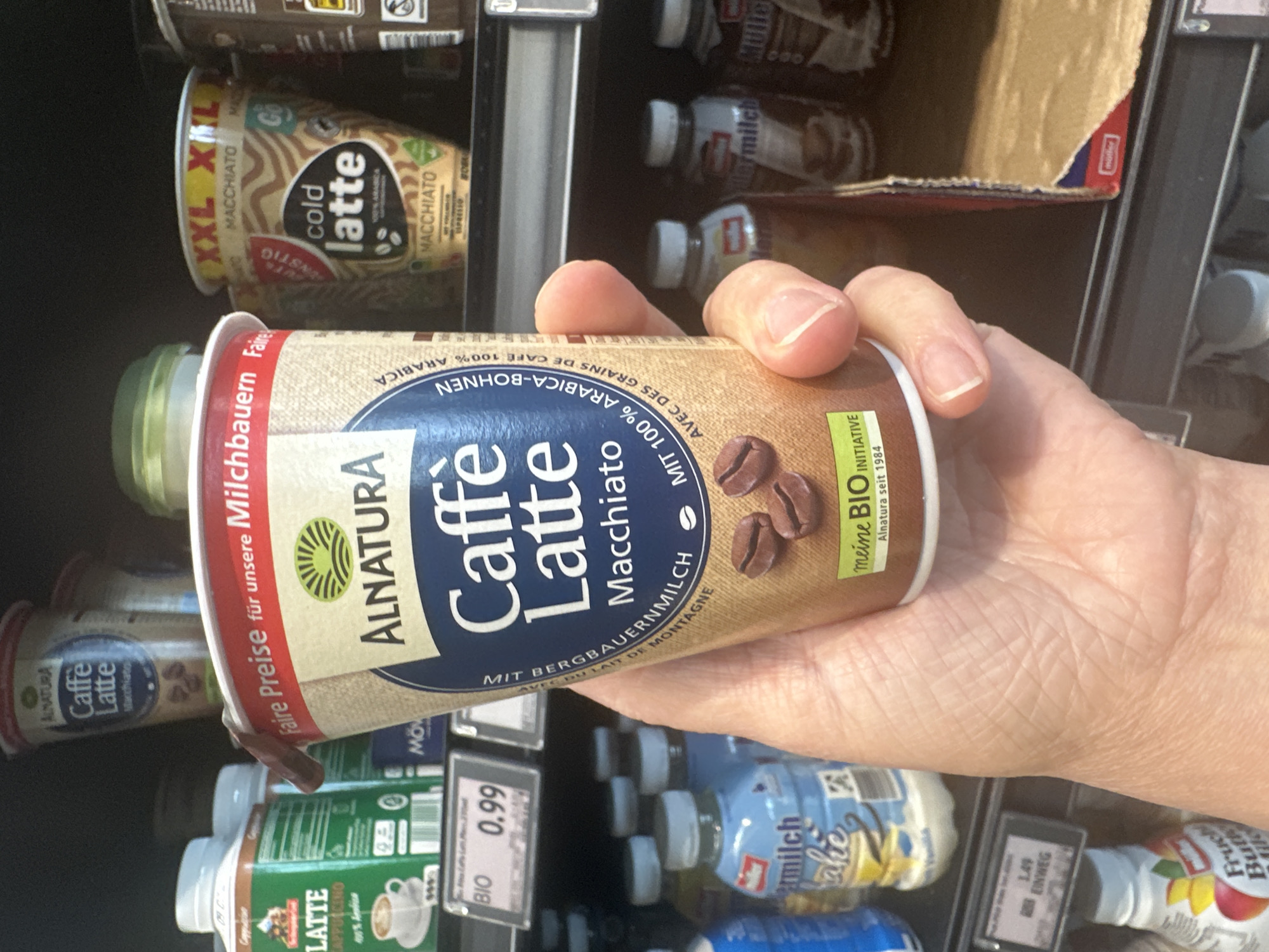 The image shows a hand holding a cup of Alnatura Caffé Latte Macchiato. The cup is beige with blue and brown text and graphics, including images of coffee beans. The background consists of shelves in a grocery store, stocked with various food and beverage items, including other coffee drinks and milk-based products. Some of the visible labels include "Alnatura", "XXL", "M Hijo", "Milch", and "Bio". The price tag "0.99" is visible on a shelf in the lower left corner. The scene appears to be in a supermarket, possibly in Tuntenhausen, Germany, given the presence of German text on the product packaging. The lighting is artificial, consistent with indoor retail lighting. The focus is on the Alnatura cup, with the background slightly blurred.