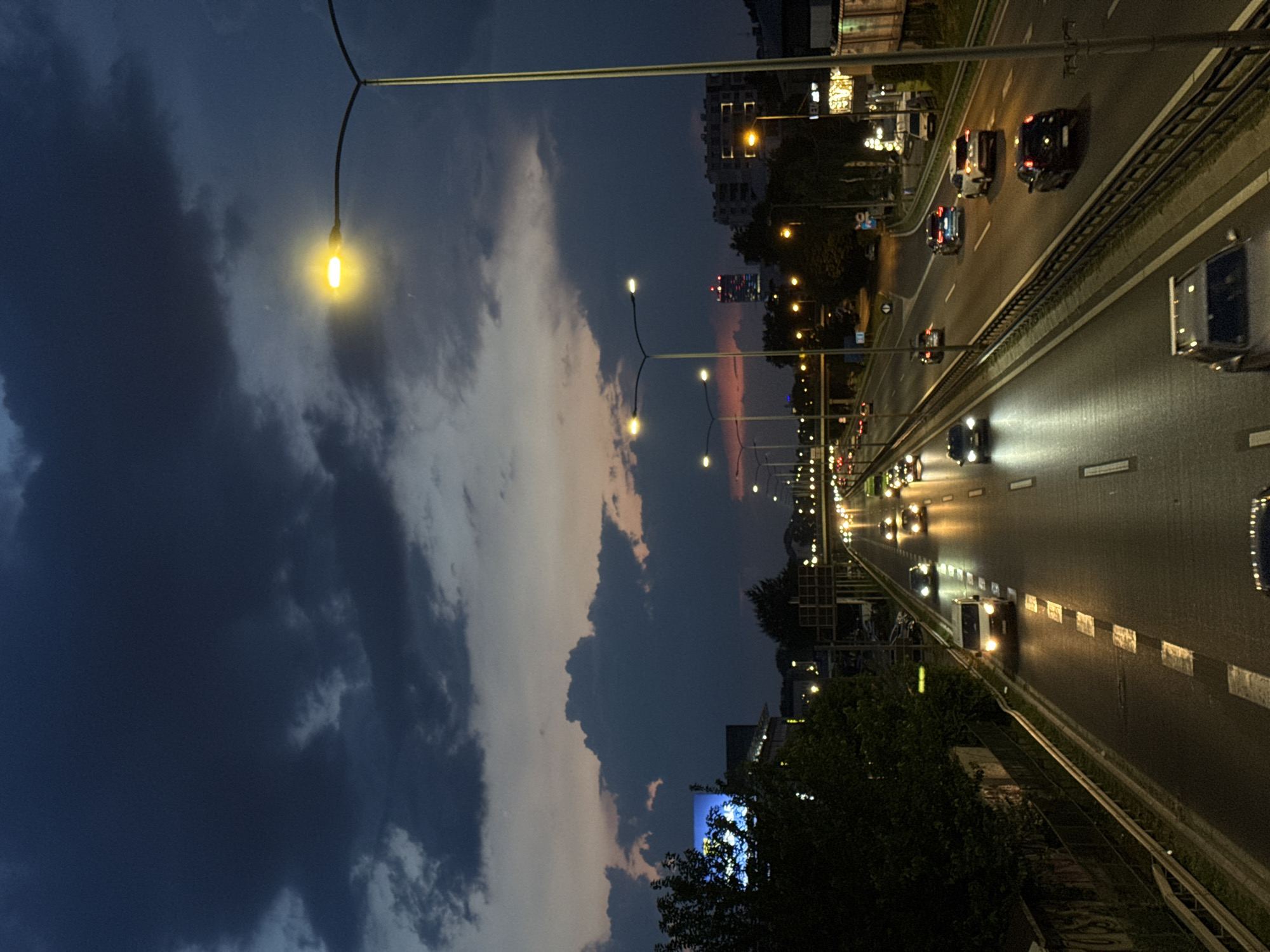 The image captures a view of a multi-lane highway at dusk, likely within the Vozdovac Urban Municipality in Serbia, given the context. The sky is dark with dramatic, heavy clouds tinged with purple and orange from the setting sun. Numerous streetlights illuminate the highway, casting a warm glow on the wet asphalt, suggesting recent rain. Cars with their headlights on are driving in both directions, creating streaks of light. On the left side of the highway, trees and a dark, graffiti-covered wall are visible, with a large, brightly lit billboard in the distance. On the right, more streetlights line the road, and buildings and structures are dimly lit in the background. A few people are visible inside some of the vehicles, but no individuals are clearly distinguishable. The overall scene is one of an active urban thoroughfare during twilight.