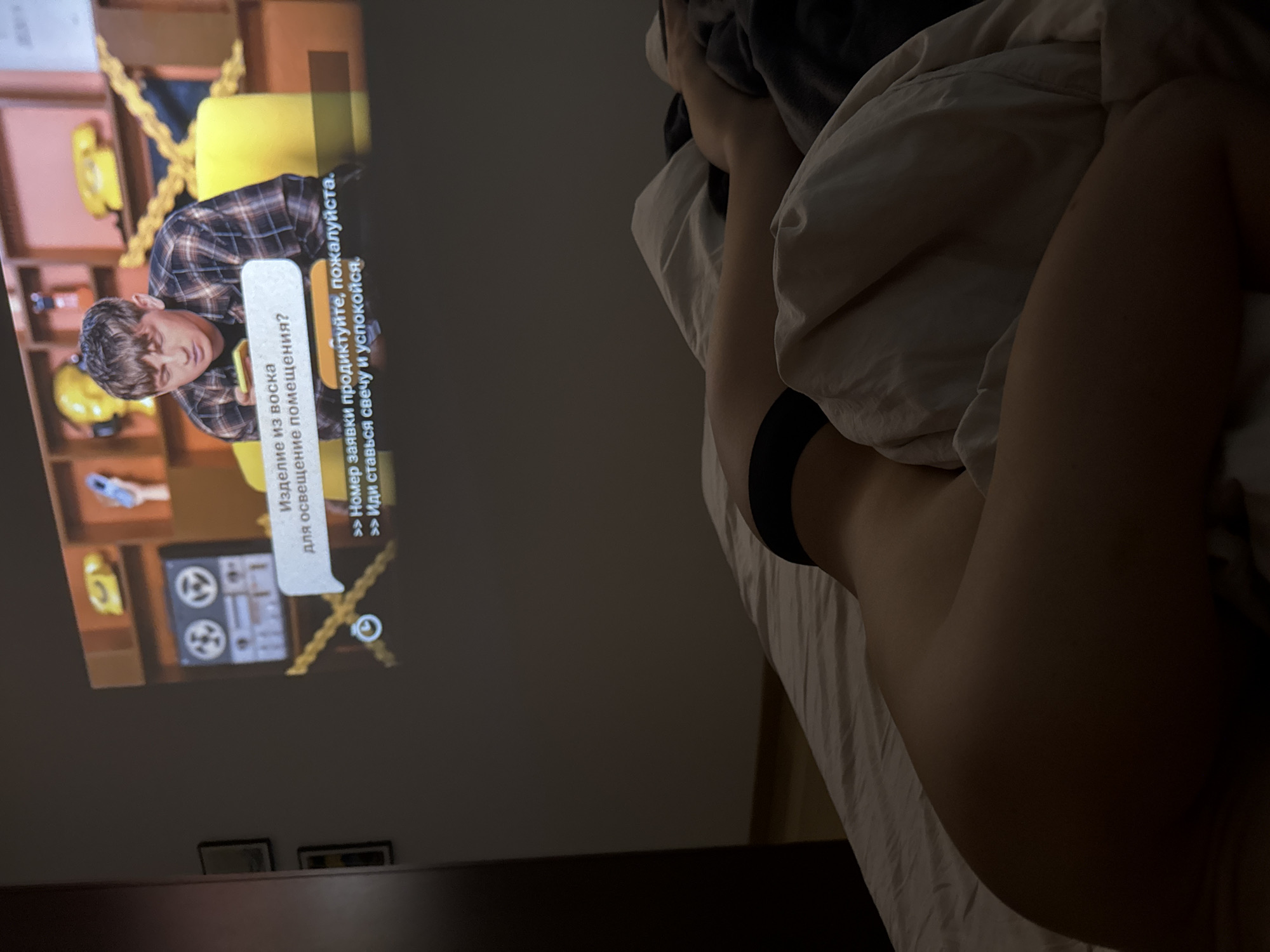 A dimly lit indoor scene features an adult laying in bed, partially covered by a light-colored duvet. The person is wearing black underwear. A projector displays a television screen on the wall. The screen shows a man wearing a plaid shirt and an on-screen menu in Russian with a question about wax decorations and options related to placing an order or lighting a candle. The overall scene suggests a relaxed or intimate setting with an individual watching television in bed. There is no data to suggest this photograph was taken in Warsaw, Poland.