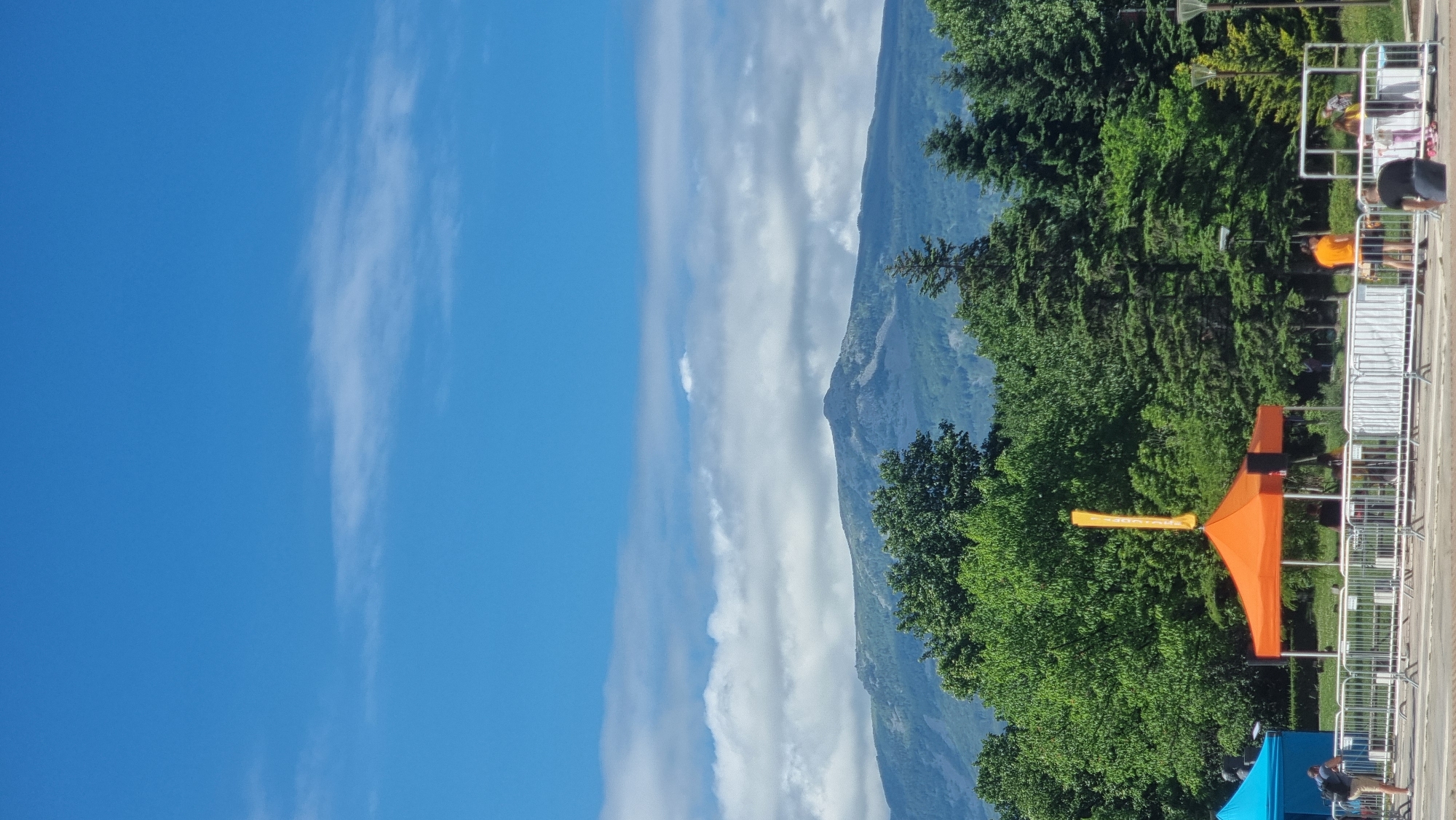 This is a vertical, eye-level shot of Vitosha Mountain, seen from the city of Sofia, Bulgaria, on a clear day with scattered clouds. In the foreground, a blue tent is visible on the left, and an orange tent with a yellow banner stands in the center, implying an outdoor event or market. Several people are present near the tents, some interacting with equipment like speakers. Metal crowd barriers are set up in front of the tents. The midground is dominated by lush green trees, forming a dense forest that extends up the lower slopes of Vitosha Mountain. The mountain itself is the dominant feature in the background, its rocky peak clearly visible against the bright blue sky, with wispy white clouds scattered across the upper portion of the frame. The overall scene suggests a pleasant outdoor setting in Sofia, with the natural beauty of Vitosha Mountain as a backdrop.