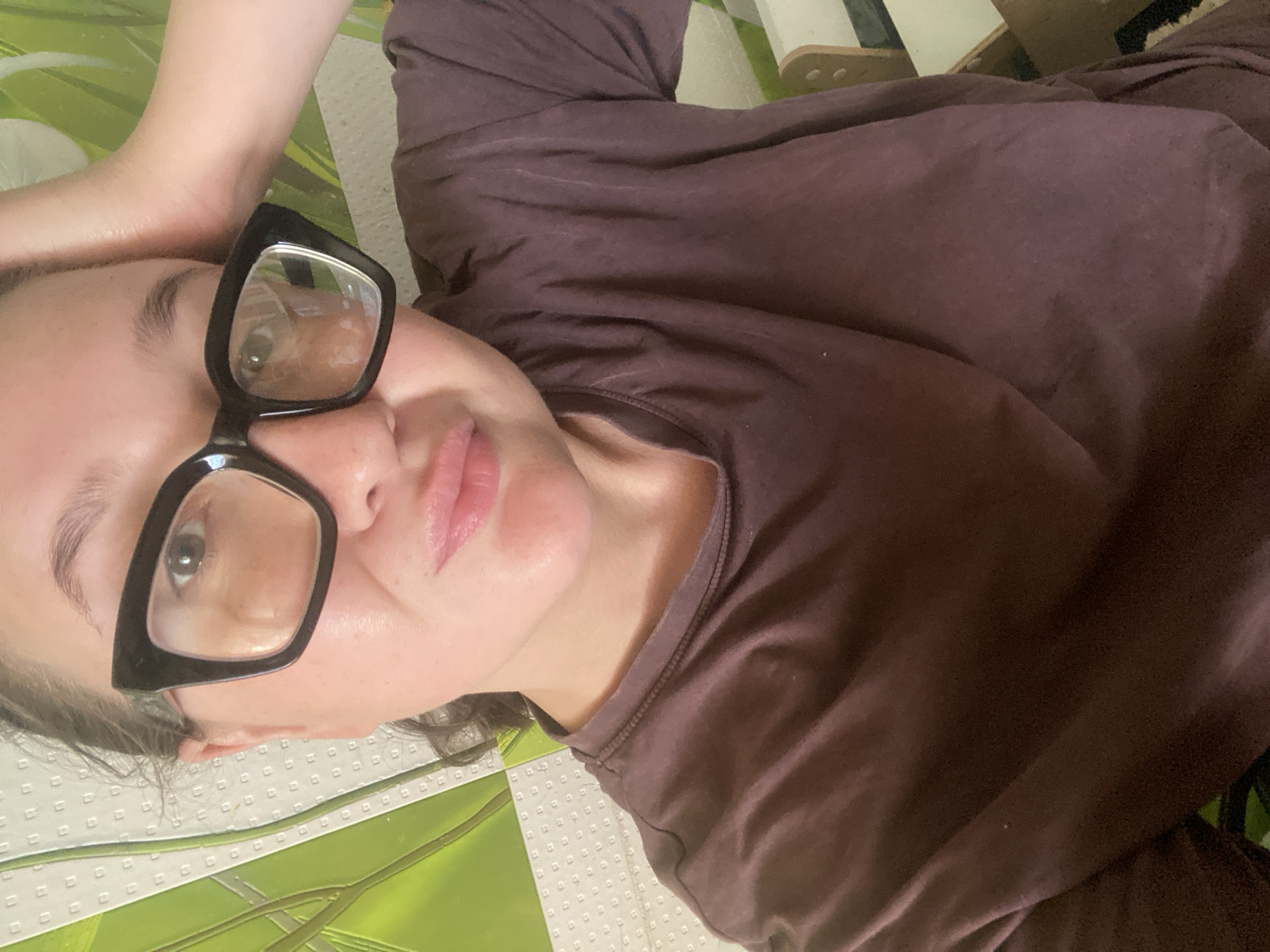 A woman with fair skin, wearing a brown t-shirt and large black-rimmed glasses, is lying on her back on what appears to be a patterned surface of white, green, and black. Her left arm is bent and placed above her head. The background includes a glimpse of a white, wooden structure, possibly part of furniture. The location is assumed to be Chaikovsky, Russia.