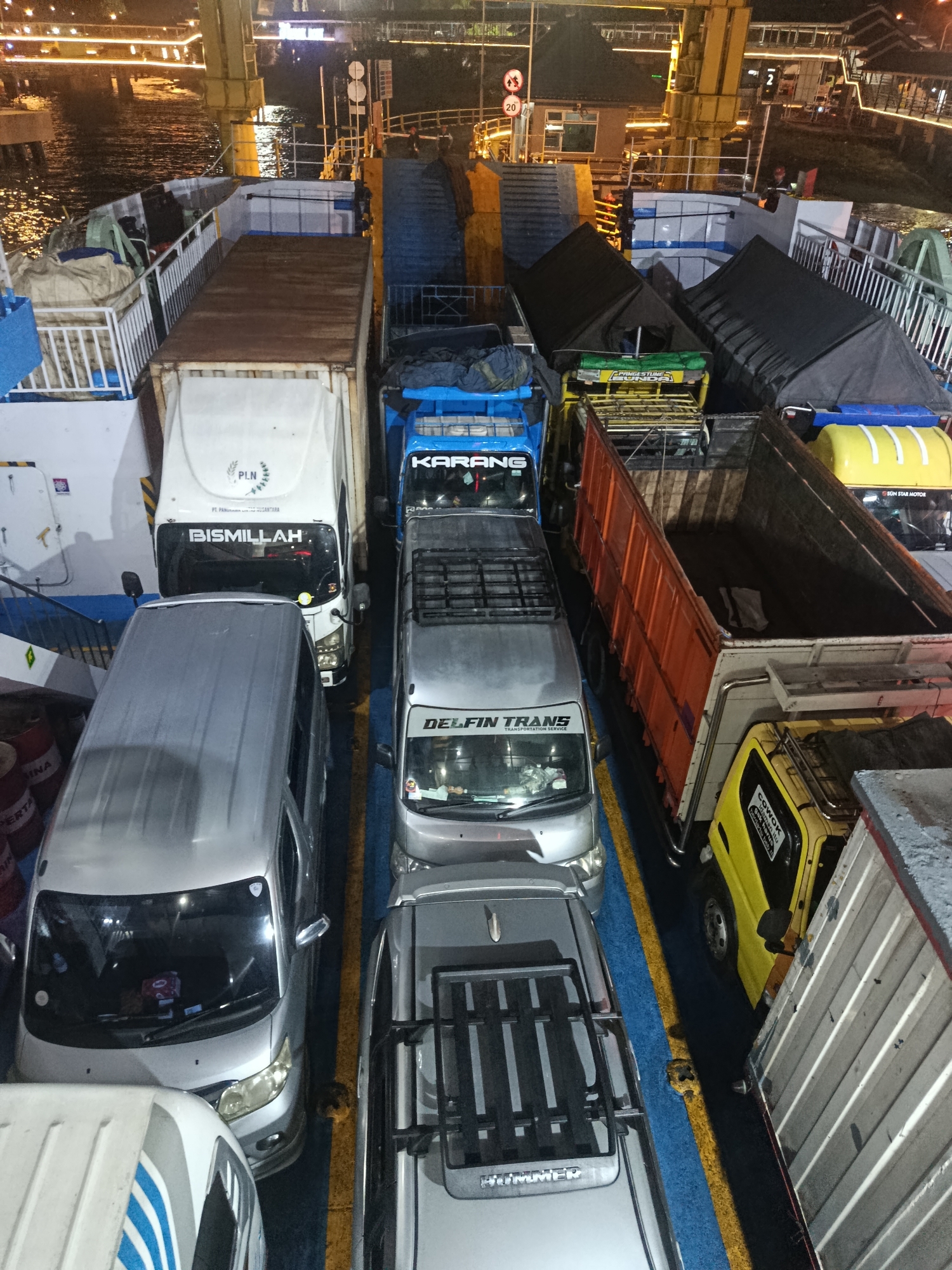 The image depicts a ferry loaded with vehicles at night. In the foreground, several cars and trucks are parked tightly together on the ferry deck. A silver van with a roof rack is positioned in the lower left, partially obscuring a white van with blue stripes. To the right of the silver van is another silver car, followed by a silver truck. Further down the line, a blue truck with "KARANG" visible on its side is parked. An orange container truck with an open back is positioned to the right, revealing its interior. A yellow truck is visible to the right of the orange truck, and a white trailer truck is at the far right. The ferry deck is marked with blue and yellow lines, indicating parking spaces. In the background, a dimly lit bridge structure can be seen, with lights reflecting on the water below. The scene is characteristic of a ferry terminal in Melaya, Indonesia, where vehicles are transported across water bodies, often at night to facilitate travel and commerce. The presence of various types of vehicles suggests a commercial ferry service.