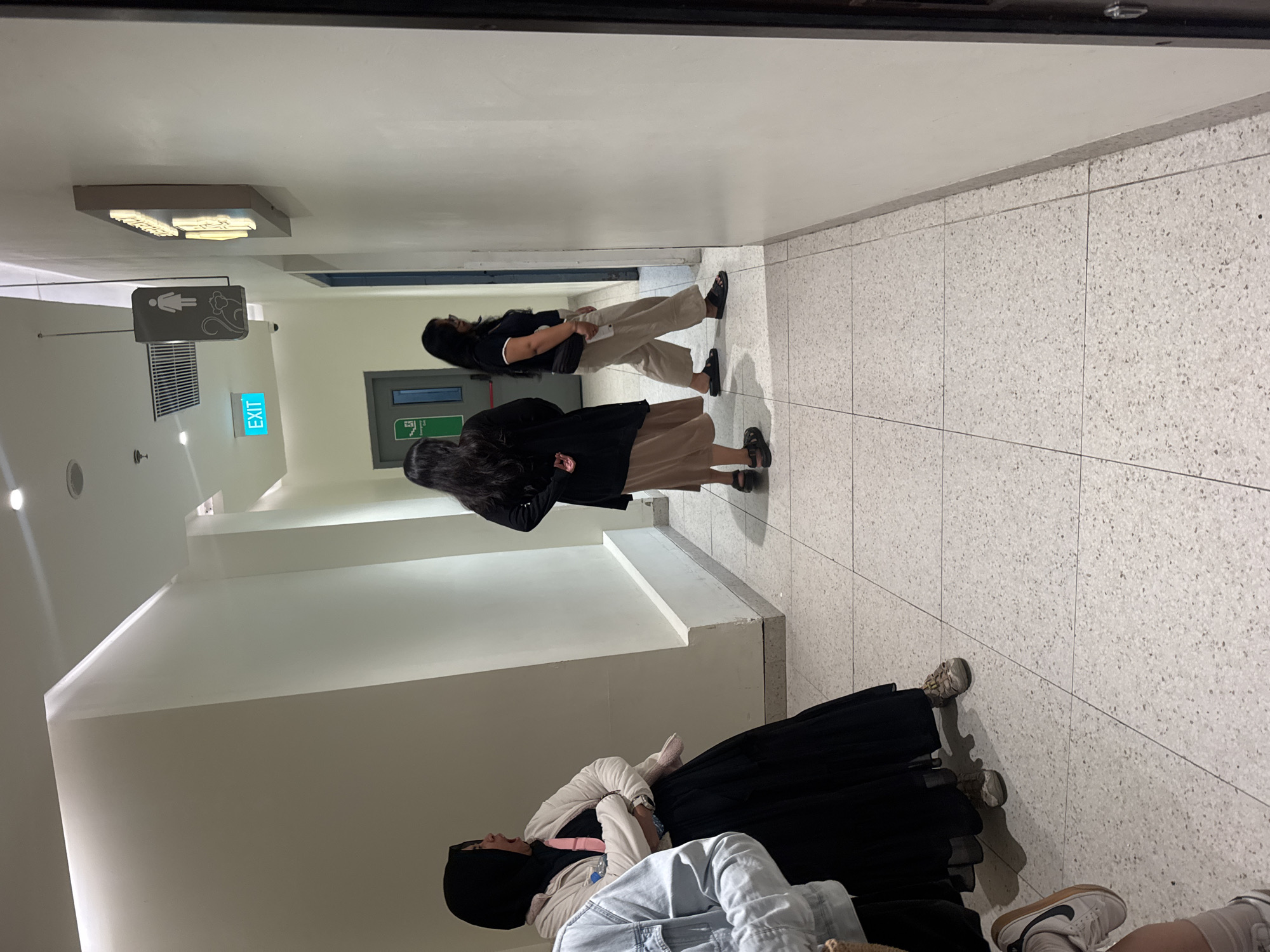 In a brightly lit, tiled hallway in Denpasar, Indonesia, three women are walking away from the camera. The woman on the far left wears a black hijab and a light-colored cardigan over a long black skirt. To her right, another woman with dark hair wears a black top and a long, light brown skirt. The woman on the far right has dark hair and wears a dark t-shirt and loose, beige pants. She carries a black fanny pack across her chest. A sign depicting a woman and a monkey hangs from the ceiling above an "EXIT" sign. A door with an "EMERGENCY EXIT" sign is visible further down the hall. The walls are a neutral, off-white color, and the floor is a speckled gray tile. The overall impression is that of a clean, modern public space.