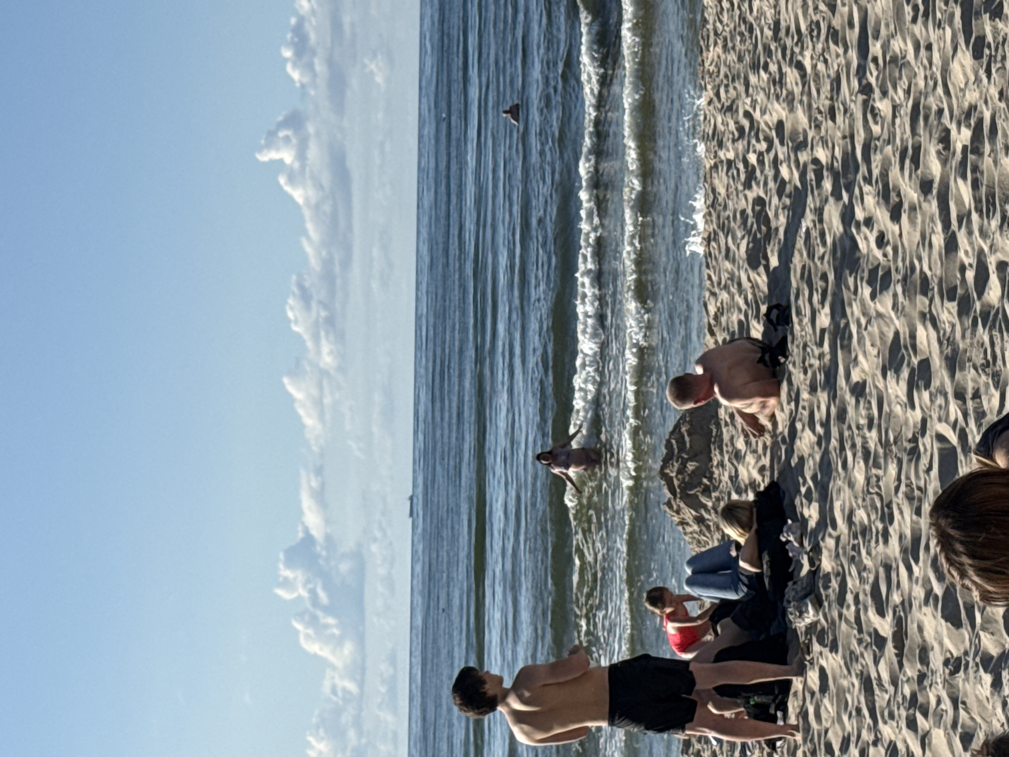 A group of people are on a sandy beach in Międzyzdroje, Poland, with the Baltic Sea in the background. The sky is blue with scattered white clouds, and a ship is visible on the horizon. In the foreground, a young man stands facing away from the camera, wearing black shorts. To his left, a young girl in a red swimsuit is crouched down, and an adult woman wearing blue jeans is lying on the sand. To the right, an adult man is lying on his back. In the sea, a woman is wading, and further out, another person is swimming. The waves are small and breaking on the shore. The sand is textured with footprints and shadows.