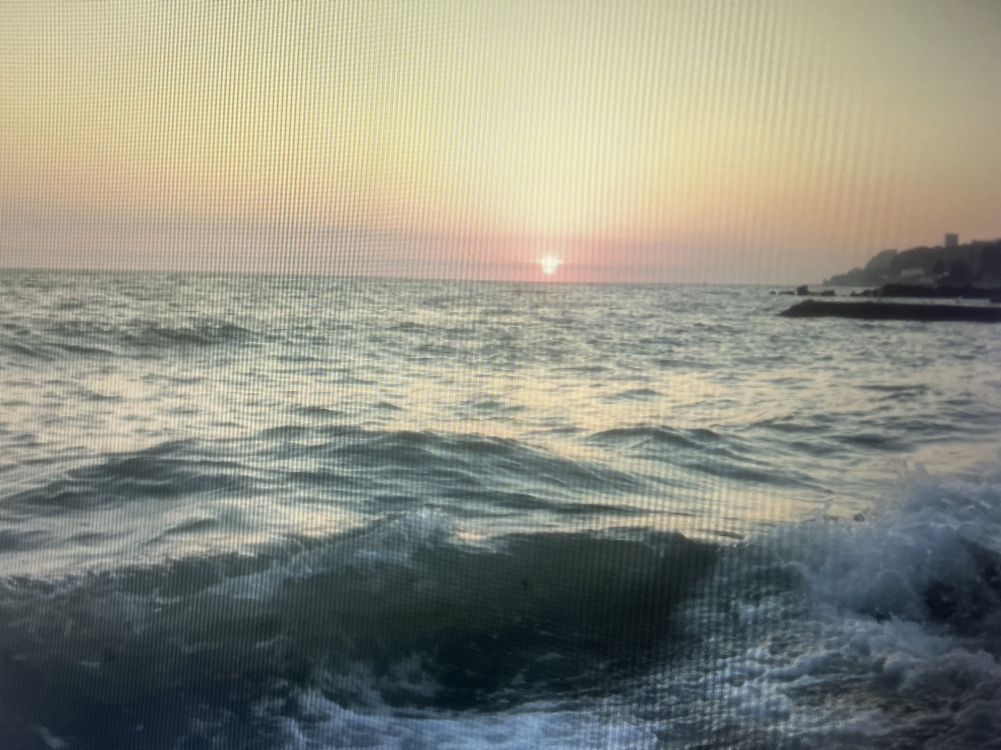 This media file captures a serene ocean scene during what appears to be sunset or sunrise. The dominant subjects are the vast expanse of the sea, with gentle waves rolling towards the viewer, and the sky, which is painted in soft hues of yellow, orange, and pink. In the distance, a coastline with silhouetted buildings and rocky outcrops is visible, suggesting a coastal town or city.
The activity taking place is the natural movement of the ocean, with waves cresting and breaking, creating white foam in the foreground. The sun, a small bright orb, is visible low on the horizon, casting a soft light across the sky and water. The overall mood conveyed is peaceful and tranquil, amplified by the soft light and the rhythmic motion of the sea.
Notable details include the warm, diffused lighting characteristic of dawn or dusk, and the slightly choppy texture of the water. The presence of the coastline with structures hints at a populated area, yet the focus remains on the natural elements. There is no visible text in the image. Based on the provided location context of Sochi, Russia, this scene likely depicts the Black Sea.
