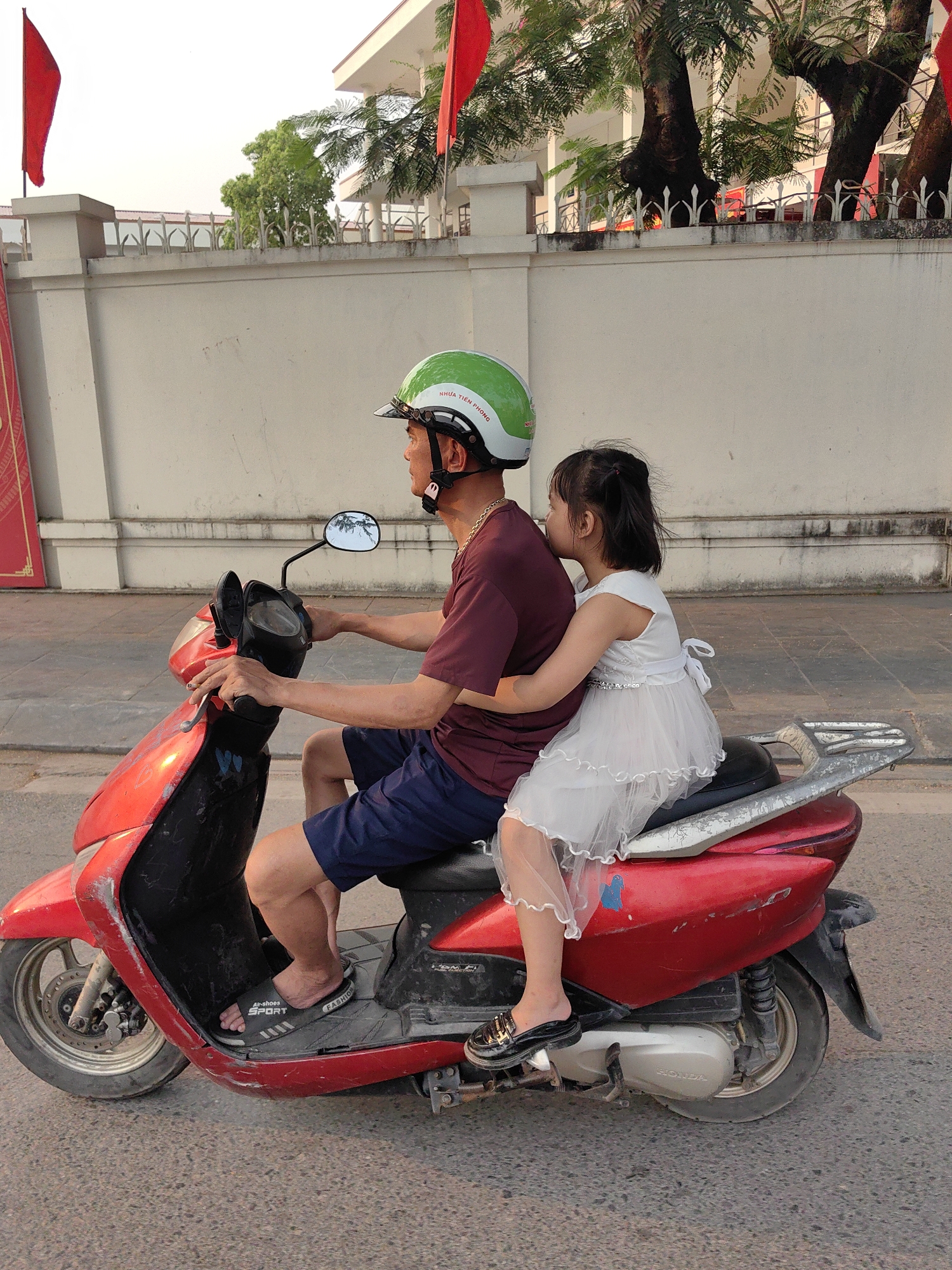 In Ha Lam Ward, Vietnam, a man rides a red Honda scooter along a city street. He wears a maroon t-shirt, blue shorts, and a green helmet. A young girl, wearing a white dress and black shoes, sits behind him, holding onto his waist. The street is paved, and a white wall with decorative fencing runs along the side. Red flags are visible in the background, near trees.