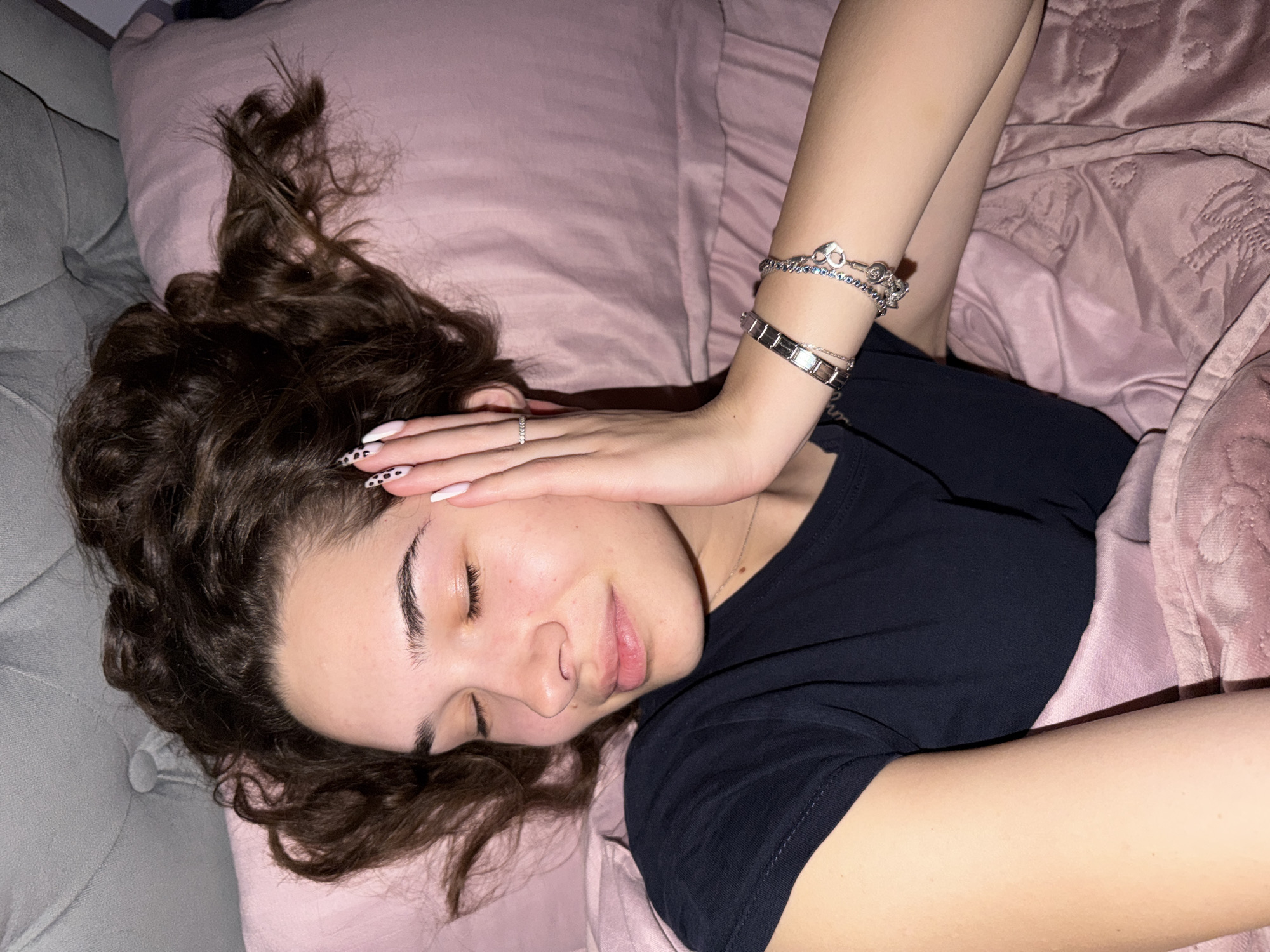 A young woman with curly brown hair is lying down on a pink pillow, with part of her head on a gray couch. She is wearing a dark blue t-shirt and several bracelets on her left wrist. Her left hand is resting on her forehead. She has a ring on her ring finger. Her eyes are closed. A pink blanket with textured squares covers part of her body. There is no evidence indicating this scene's location is in Dobrich, Bulgaria.
