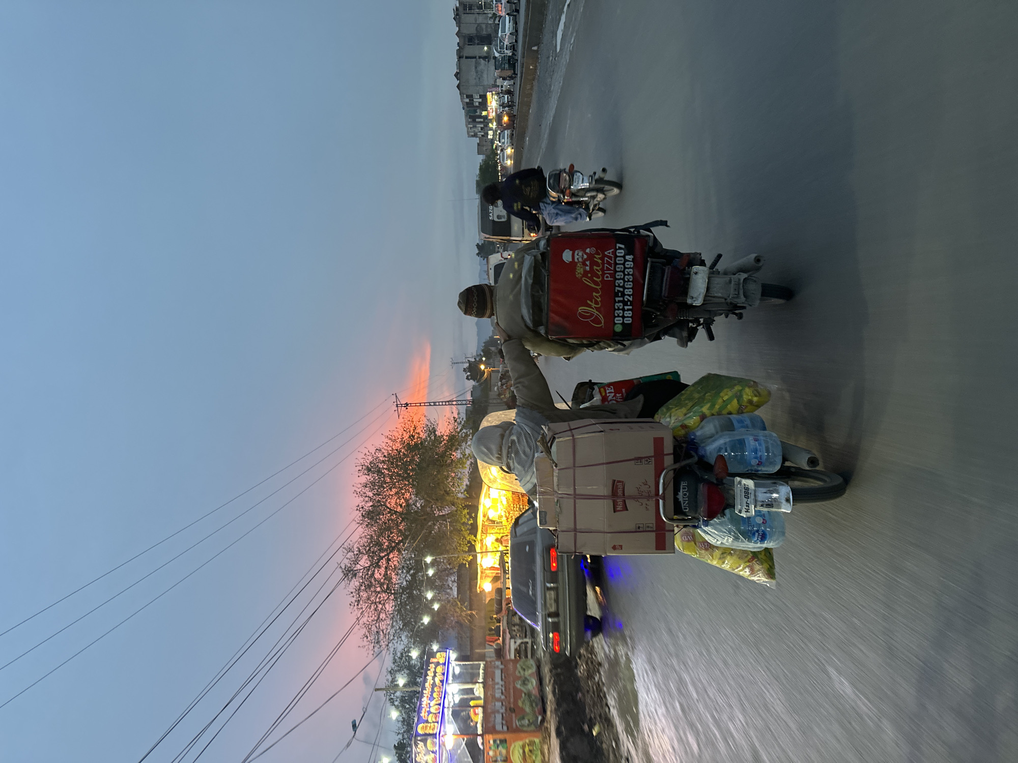 The image depicts a street scene in Quetta City Tehsil, Pakistan at dusk. The sky is a gradient of blue and orange, with buildings visible in the background. In the foreground, two motorcycles are traveling on the road. The first motorcycle is carrying a large cardboard box, bags of groceries, and several bottles of water. The second motorcycle behind it is carrying a pizza delivery box with the words "Italian Pizza" and a phone number on it. The road appears to be wet or in motion, suggesting that the photo was taken from a moving vehicle. Along the side of the road are lights and stalls, likely part of a commercial area. Power lines are visible in the upper left of the frame.