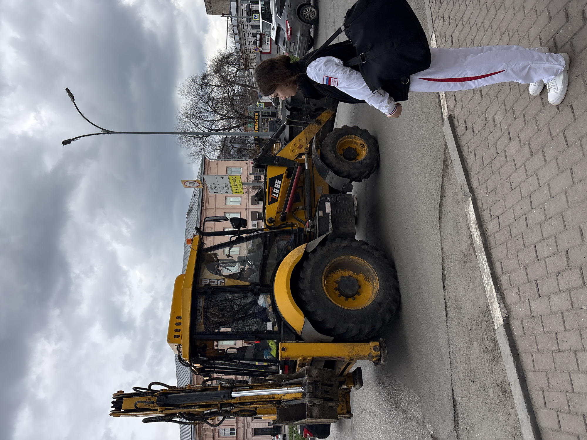 A woman in a white tracksuit with a red stripe down the side and a black backpack is standing near a yellow JCB LB 95 backhoe loader on a paved street. The background includes buildings, cars, a street sign with Russian writing that includes references to "Ulitsa" and "Lesnika" (street and forester), and a streetlight. The sky is cloudy. This scene is likely in Ulyanovsk, Russia.