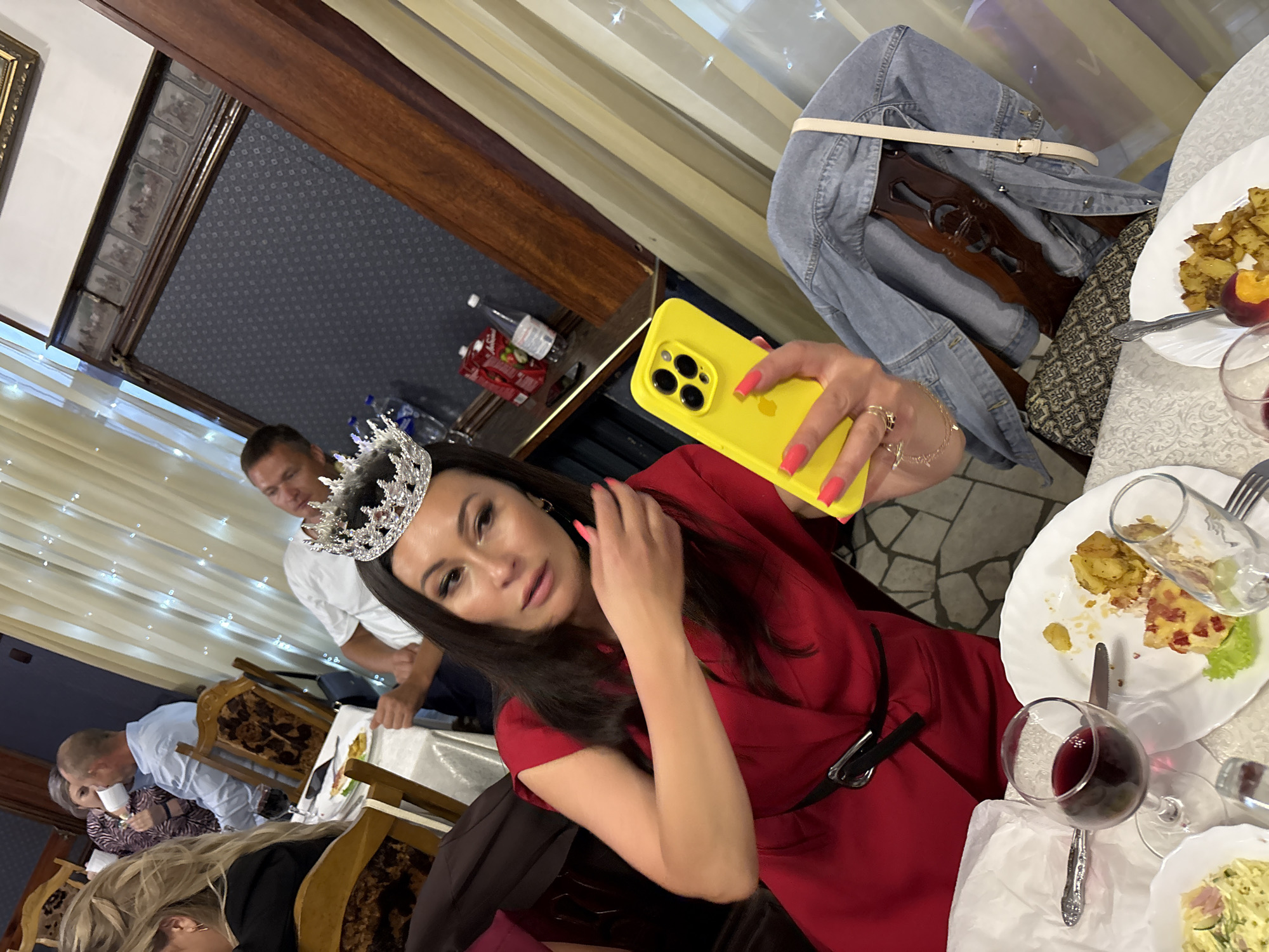 This media file captures a group of people gathered around a table, likely at a celebratory event or a meal. In the foreground, a woman wearing a sparkling tiara and a vibrant red dress is taking a selfie with a yellow smartphone. Her nails are painted a bright coral color. To her left, a man in a white shirt is visible, also appearing to be part of the gathering.
The setting appears to be indoors, possibly a restaurant or a banquet hall, suggested by the tablecloths, chairs with ornate carvings, and decorative lighting in the background. The table is laden with plates of food, including what looks like potatoes and a pastry, as well as glasses of red wine. A denim jacket is draped over the back of a chair.
The lighting suggests it could be evening. The overall mood seems festive and convivial, with the woman in the tiara perhaps being the center of attention, possibly a pageant winner or celebrating a birthday. The presence of multiple people and the dining setup indicate a social gathering. The location context provided is Zavod Mikhailovskii, Russia. No visible text is present in the image.