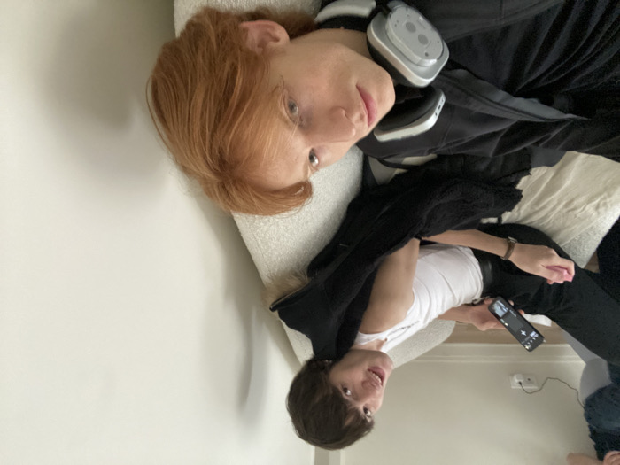 The image displays an indoor residential scene featuring two individuals reclining on a light-colored, textured sofa against a plain white wall. No visual cues within the image definitively confirm the location as Paris, France; however, the scene depicts an indoor setting.
In the upper-left and center of the frame, a male individual with dark brown hair, wearing a white sleeveless top and dark trousers, is positioned with his torso angled towards the viewer. He holds a black smartphone in his left hand, displaying a user interface, and a small pink object in his right hand. A brown wristwatch is visible on his left wrist. He is looking directly at the camera with an open-mouthed expression. A black garment, possibly a jacket, is draped over his lap and the sofa cushion. A white electrical outlet with a plugged-in white cable is visible on the wall behind him.
In the foreground, another male individual with reddish-brown hair is lying or reclining on the sofa, oriented perpendicular to the first person. He is wearing a dark, possibly hooded, garment. A pair of light grey and white over-ear headphones rests around his neck. He is looking towards the camera with a neutral expression. The interaction between the two individuals is primarily their shared orientation towards the camera. The environment suggests a casual, relaxed atmosphere.