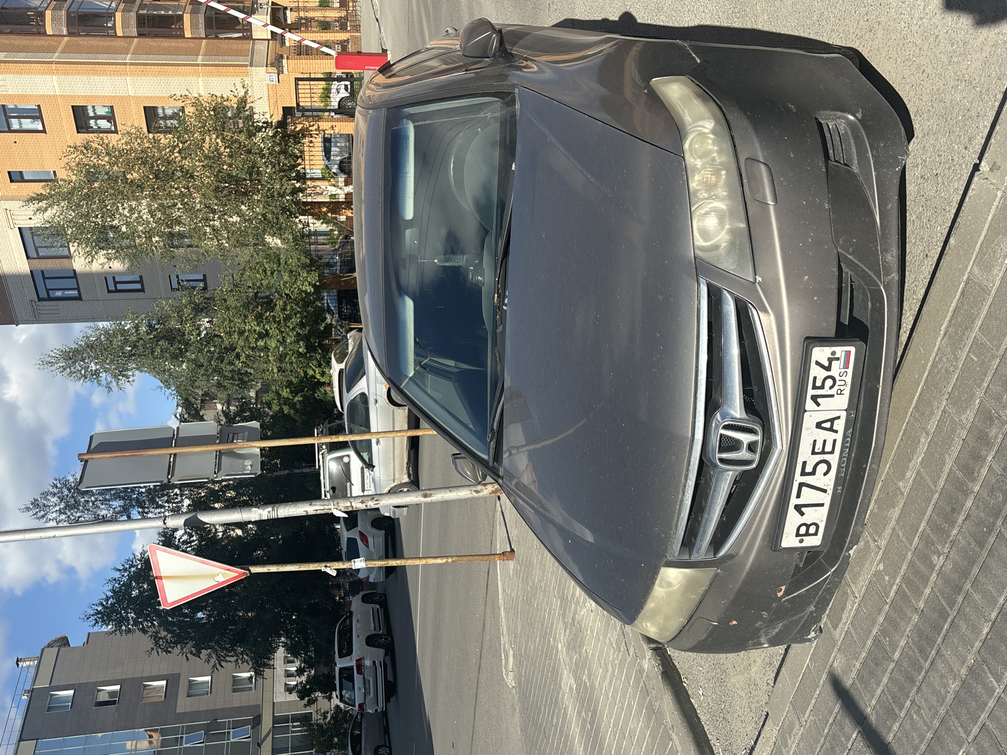 The image shows a gray Honda sedan parked on a paved street in Novosibirsk, Russia. The car is facing the viewer, and its license plate reads "B175EA 154 RUS". The car appears to be slightly damaged, with scuff marks on the front bumper. In the background, there are several buildings, including a modern apartment complex and an older brick building. There are also trees and street signs visible. The sky is blue with some clouds. The scene appears to be a typical urban environment on a sunny day.