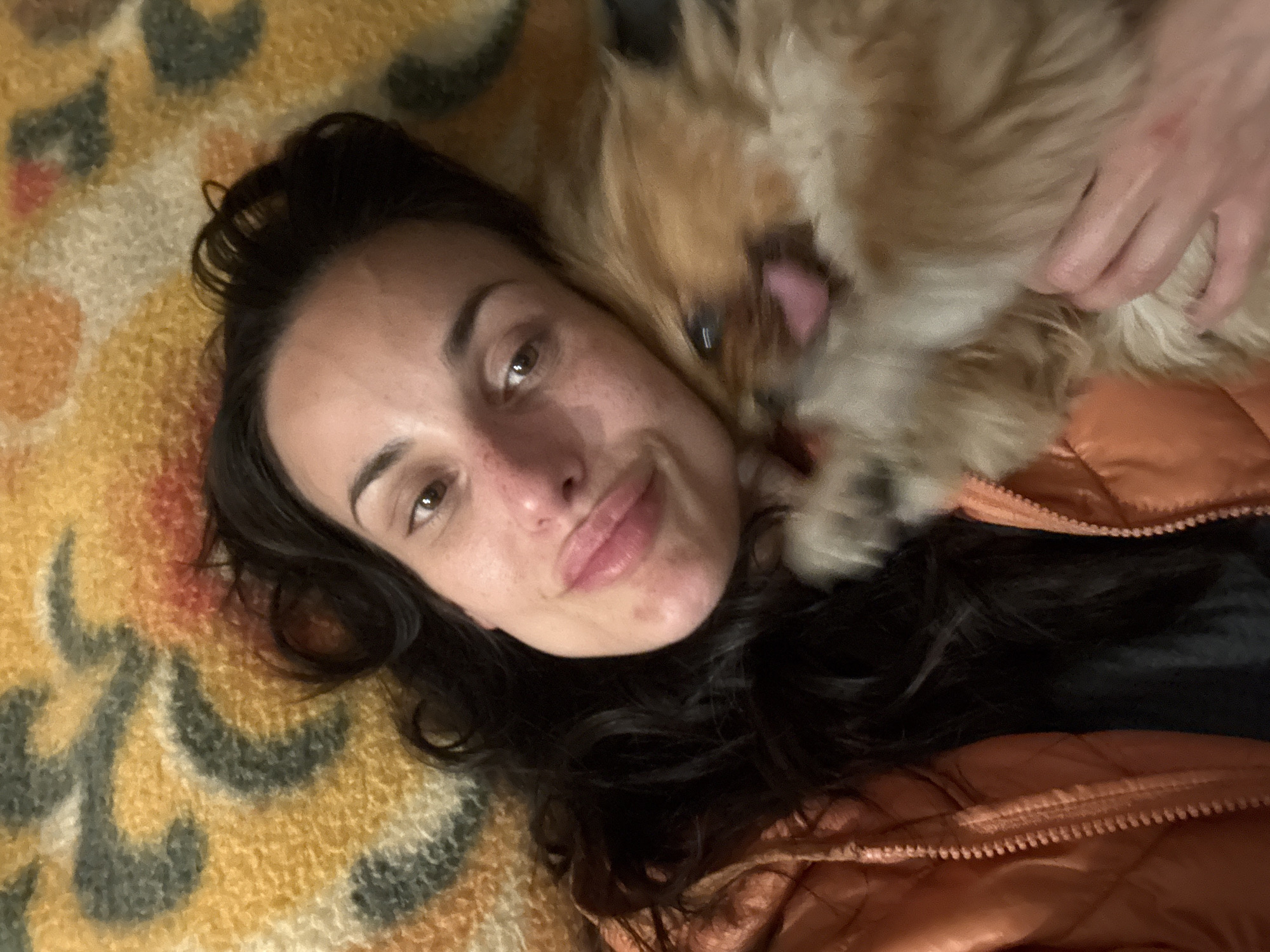 A woman with dark hair is lying down on a yellow and orange patterned blanket. A small, fluffy, tan-colored dog is lying next to her, licking her face. The woman is wearing a copper-colored quilted jacket. The location is unknown and there is no information in the image to confirm the scene is in Pleven, Bulgaria.