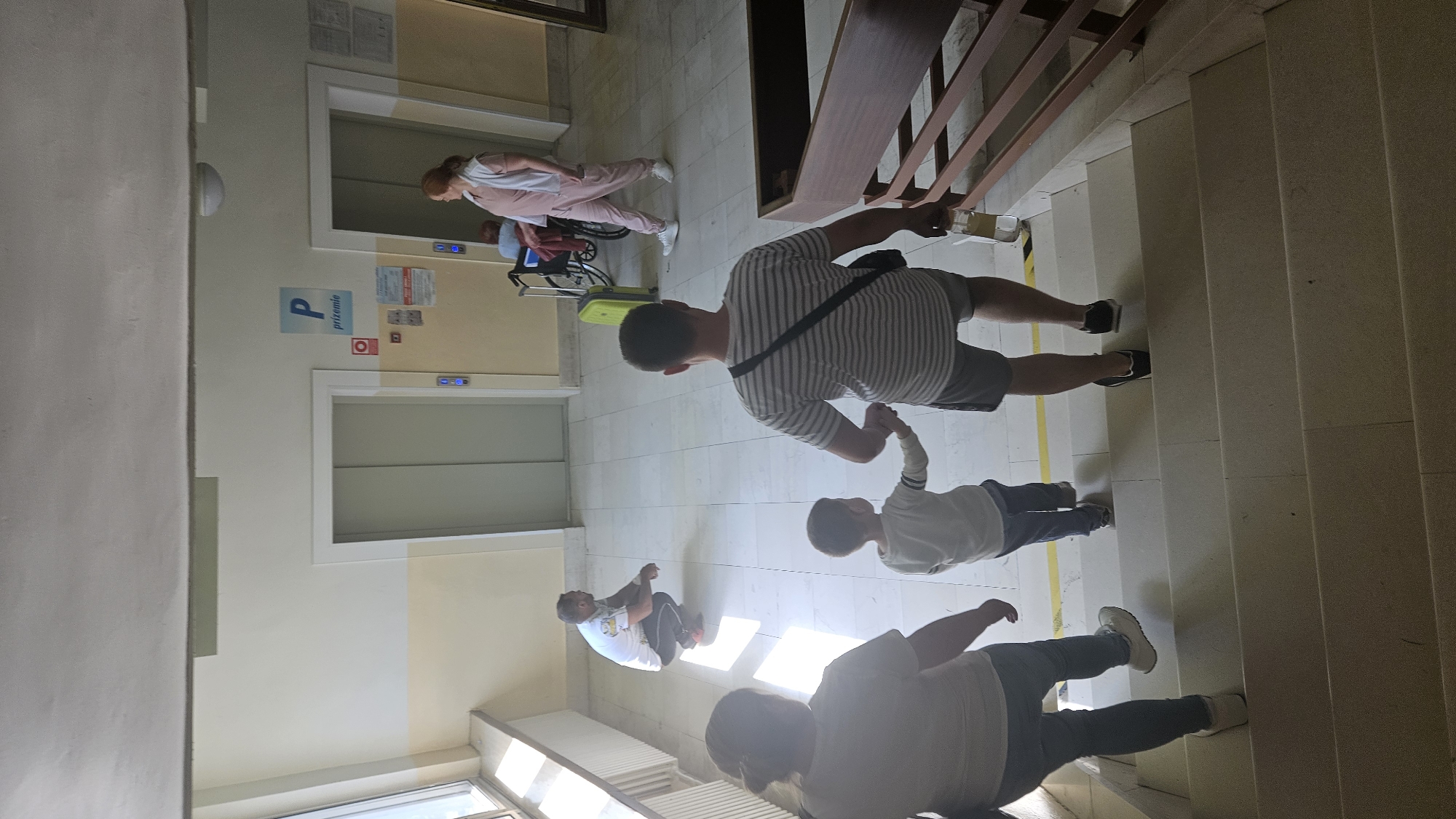 The image depicts a family descending a staircase within a building that appears to be a medical facility or a public institution in Prešov, Slovakia, judging by the "P prizemia" sign, which translates to "P ground floor" in Slovak. Two elevators are visible on the ground floor, one with its doors open. A woman in a pink outfit is standing near one of the elevators, and a person in a wheelchair is visible behind her. A bright yellow suitcase is also present on the ground floor. On the staircase, a man wearing a striped shirt and shorts is holding the hand of a young boy in a light-colored shirt. Following them down the stairs are a woman in a white shirt and jeans and another person whose face is not visible. The building's interior features tiled floors, pale yellow and white walls, and a window on the left side. The overall impression is of a mundane moment of people navigating a multi-level building.