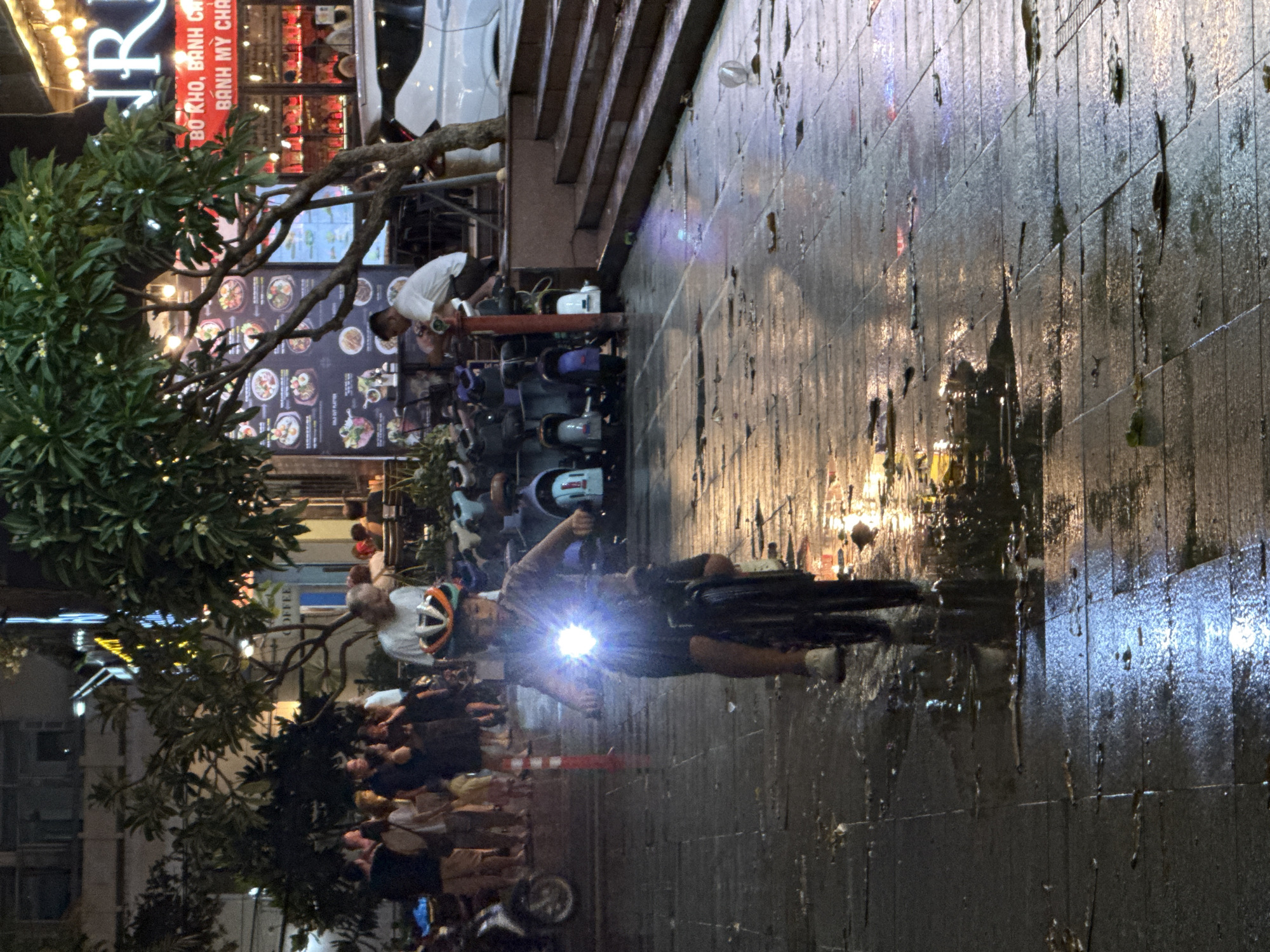 A young person wearing a helmet rides a bicycle with a bright headlamp down a wet, tiled sidewalk at night. The sidewalk is illuminated by the bright light from the bicycle's headlamp and reflections from nearby building lights. In the background, a group of people stand and talk near a restaurant with a menu displayed outside. Several scooters are parked on the sidewalk to the left of the cyclists. The scene appears to be on a street in Nha Trang, Vietnam, characterized by its vibrant nightlife and pedestrian-friendly areas. The wet conditions suggest it may have recently rained or is still raining.
