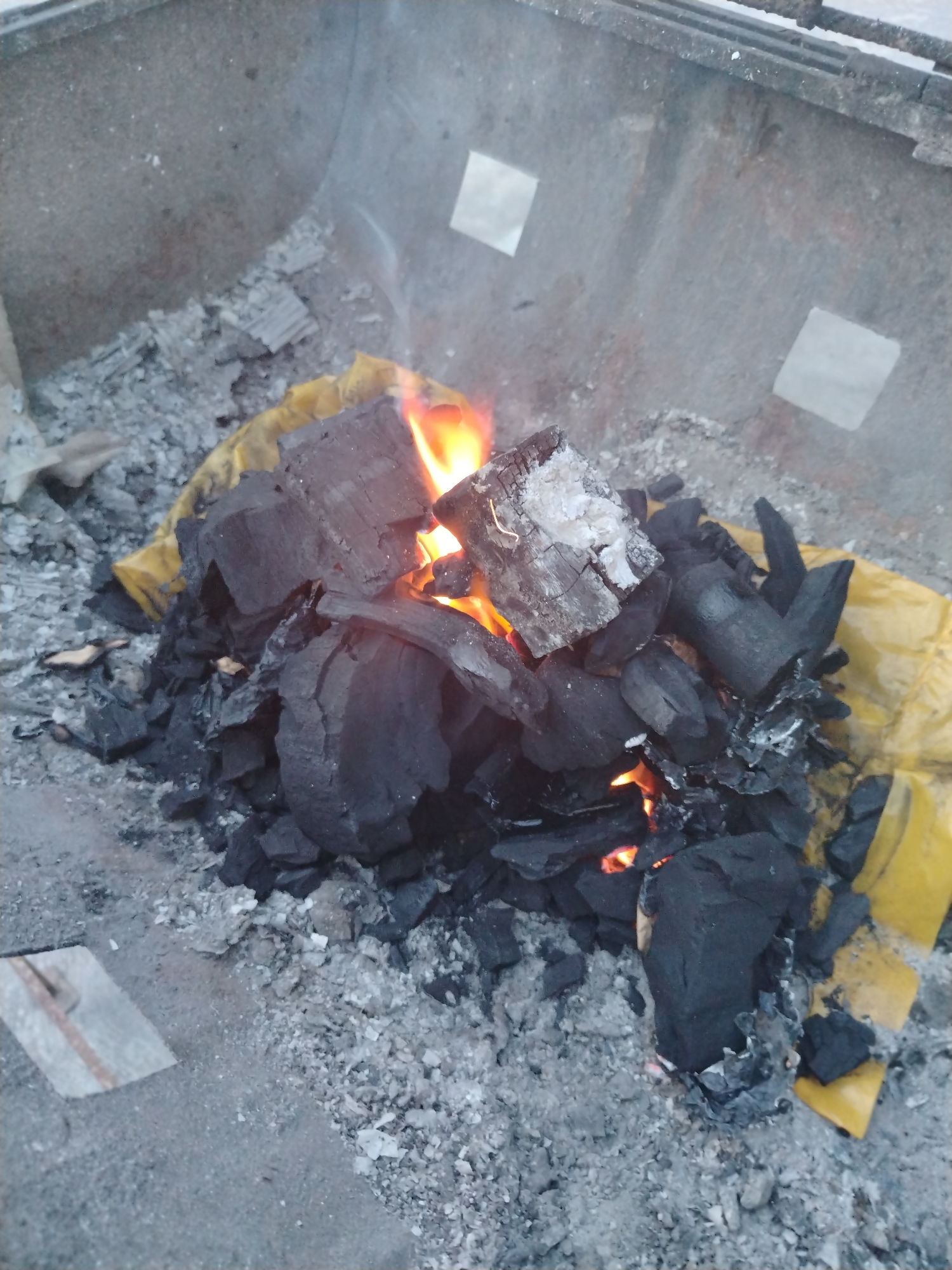The image displays a close-up view of a charcoal fire burning within a metal structure, likely a grill or a makeshift fire pit, in Tinaquillo, Venezuela. A significant pile of black charcoal pieces occupies the central area, with several chunks actively engulfed in bright orange and yellow flames, indicating that the fire is in full swing or has recently been lit. Some charcoal pieces exhibit white ash residue, suggesting they have been burning for some time.
Beneath and partially melted around the charcoal, remnants of a yellow plastic sheet or bag are visible. This material appears burnt and crinkled, likely having served as kindling or packaging for the charcoal. The base of the metal container is covered in a layer of fine grey ash and small debris. The interior walls of the metal structure are visible in the background, appearing somewhat grimy and weathered. Notably, there are small, square, light-colored or reflective patches adhered to the inside surfaces of the metal walls.
The ground surrounding the fire pit is a rough, grey surface, possibly concrete or asphalt, also scattered with ash and small fragments. The overall scene depicts the process of starting or maintaining a charcoal fire, commonly used for cooking or providing warmth. No individuals are visible, and there is no discernible text within the image. The lighting is diffuse, making the exact time of day difficult to determine, but it does not appear to be direct daylight.