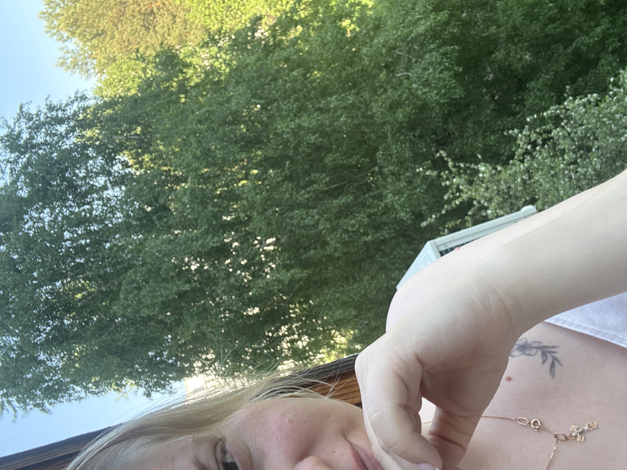 A selfie appears to have been taken from a low angle, featuring a person's face and arm in the foreground, with a background of green foliage and glimpses of sky. A building or structure is barely visible behind the person's arm. The individual has light-colored hair and is wearing a necklace with a pendant. A small tattoo is visible on the chest/shoulder area. The location cannot be determined from the visual information provided. The city of Chaikovsky, Russia, cannot be confirmed without additional context.
