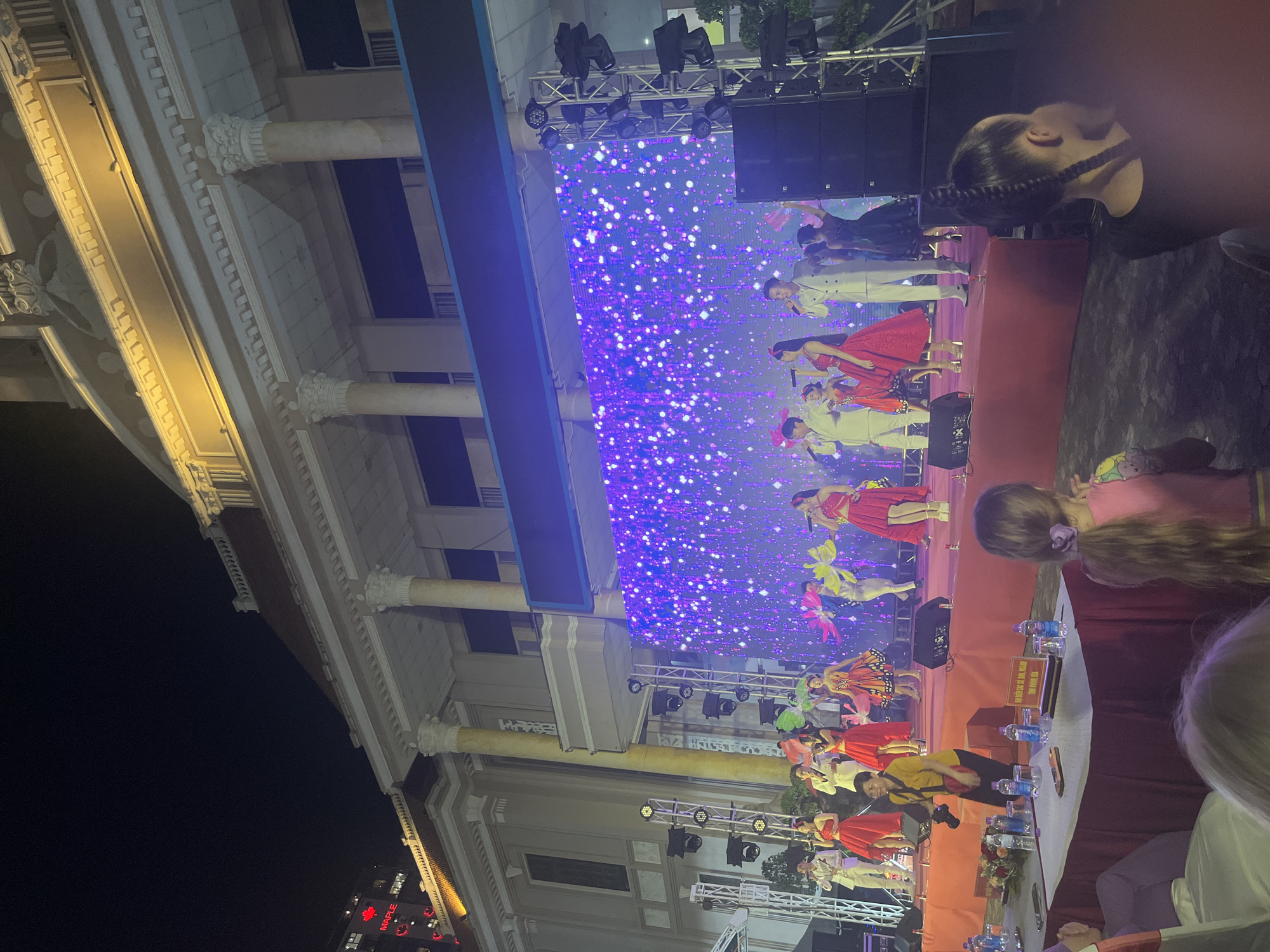 At night, an outdoor stage is set up in front of a large building with classical architecture and illuminated columns. A group of performers, including singers and dancers, are on stage. The backdrop is a large LED screen displaying a sparkling, purple, and blue pattern. The performers are dressed in colorful costumes, with some in red dresses, a woman in a butterfly-patterned dress, and men in white suits. They are holding microphones and appear to be singing and performing. In the foreground, a long table is set up with bottles of water and some floral arrangements. Spectators are seated at the table, and some individuals are standing and watching the performance. In the very foreground, partially visible, are audience members, including two girls facing away from the camera and looking towards the stage. A woman in a yellow shirt is standing on the edge of the stage holding a camera. The scene suggests a public event or performance taking place in a city.