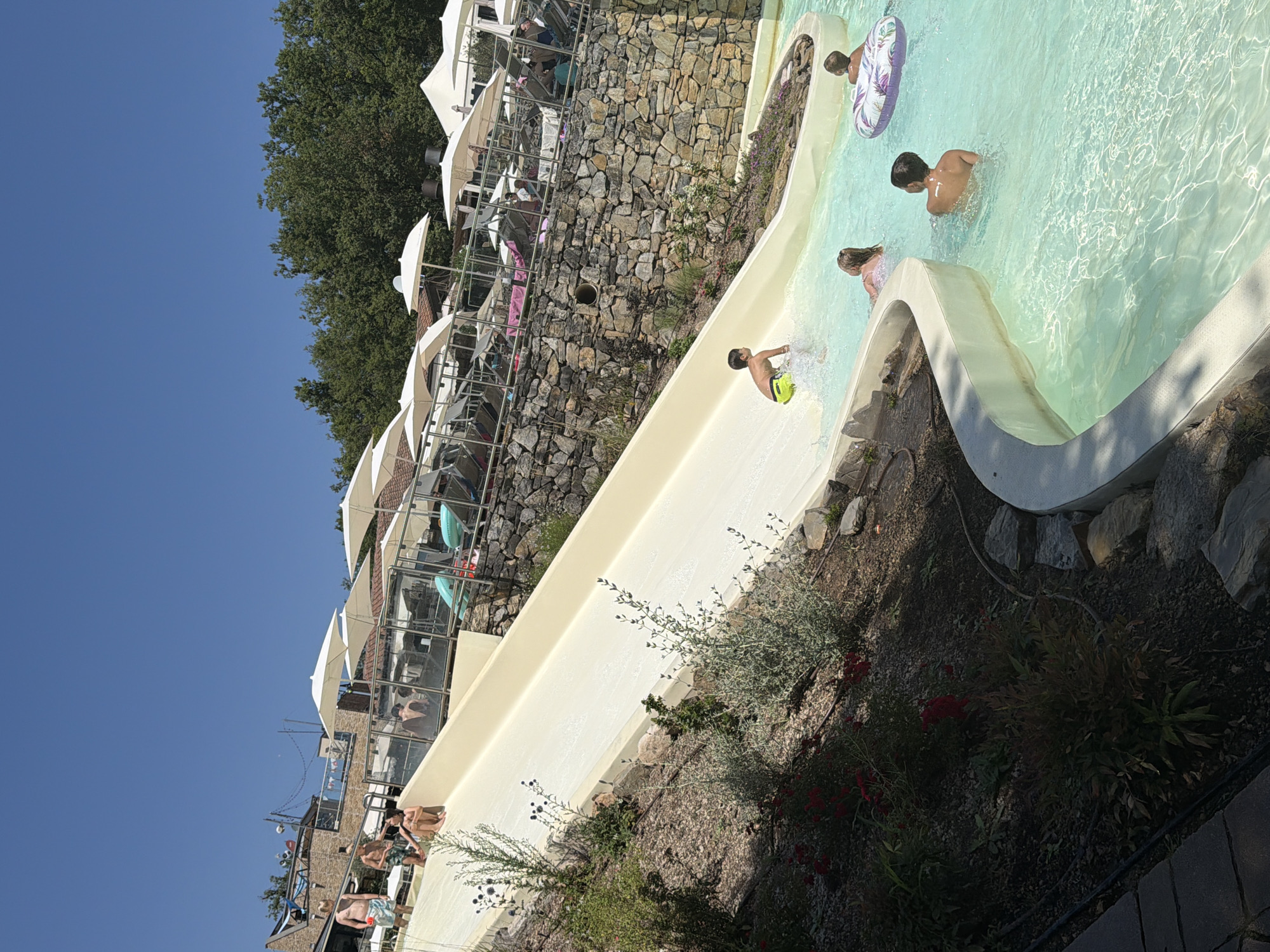 In Cavriglia, Italy, a bright, sunny day is captured at a water park. A wide, cream-colored water slide dominates the foreground, with a young boy in neon yellow swim trunks mid-slide at the bottom, splashing into a clear turquoise pool. To the right of the slide's end, a girl with blonde hair is partially submerged in the pool, and further right, a boy wearing a striped inner tube floats on his back. Another boy is visible wading in the pool. The pool area features a curved white edge, contrasting with the rocky, landscaped embankment on the left, which is dotted with green shrubs and red flowers. In the background, a stone wall supports an elevated area with lounge chairs and numerous white umbrellas providing shade. Several people are visible relaxing on the lounge chairs. The sky above is a vibrant, clear blue.