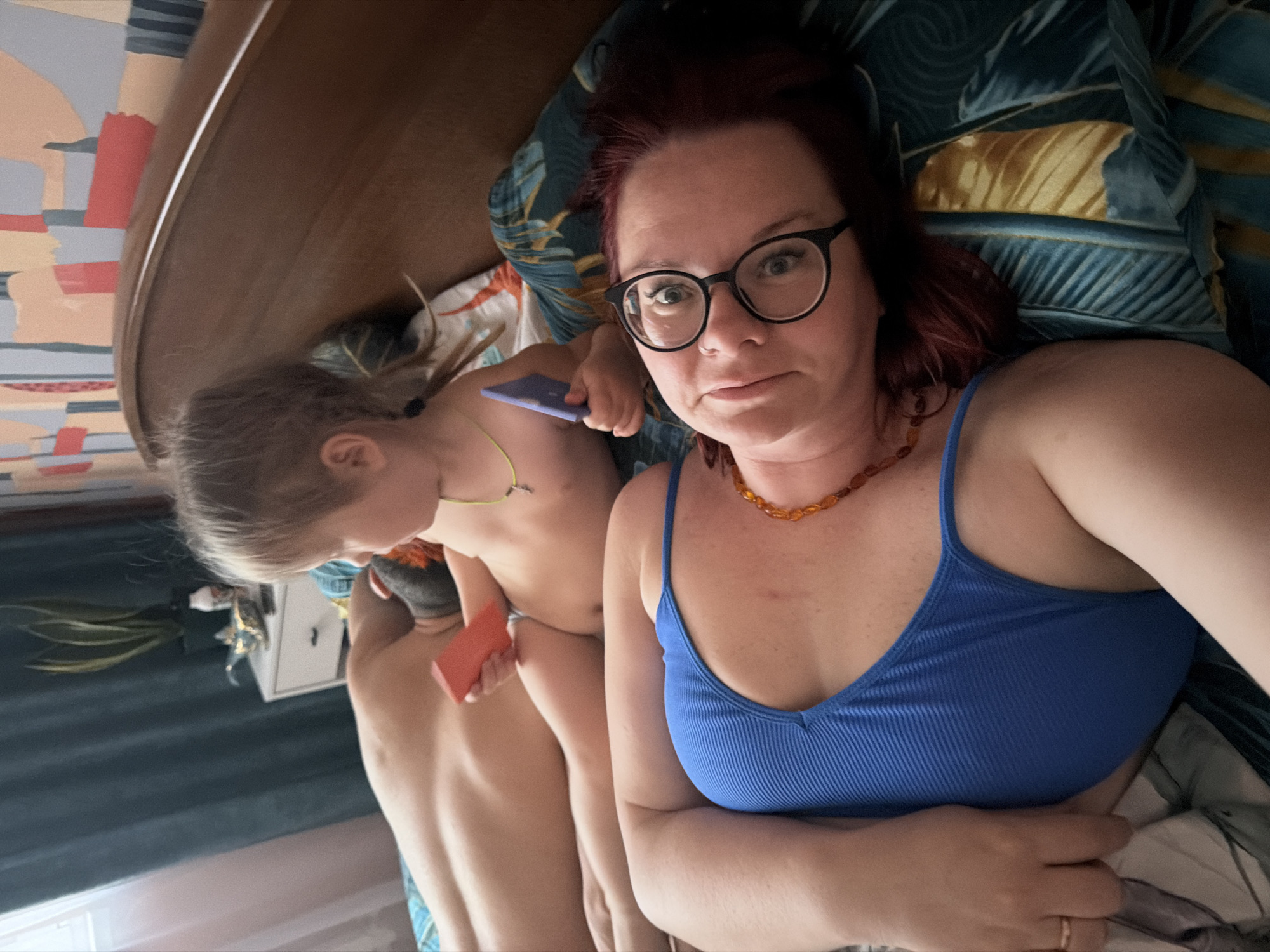 A woman wearing glasses and a blue tank top lies on a bed with patterned pillows. A child with blonde hair is next to her, holding two rectangular objects, one purple and one orange. The environment includes a wooden headboard, a wall with a decorative pattern, and a white nightstand with a plant on it. The image appears to be taken indoors. The exact location in Chaikovsky, Russia cannot be determined from the image.