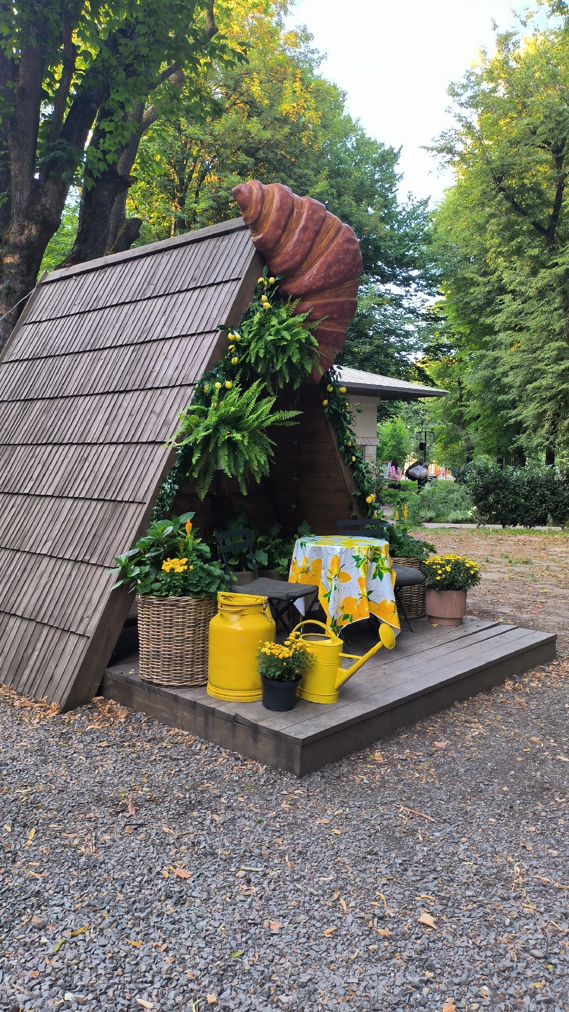 The image depicts a quaint outdoor seating area with a whimsical, oversized croissant sculpture adorning the triangular wooden structure. The structure itself resembles a stylized A-frame building with wooden shingles. Lush green ferns and trailing ivy with faux lemons are draped around the croissant and the eaves of the structure.
In front of the structure, a wooden deck serves as the seating area. A round table is covered with a yellow tablecloth featuring a lemon print. Two dark-colored chairs with woven bases are placed around the table.
On the deck, several decorative items are arranged:
- A large, woven basket contains yellow flowers.
- A bright yellow, vintage-style milk can stands next to the basket.
- A small black pot holds more yellow flowers.
- A yellow watering can with a long spout is positioned on the deck.
- A terracotta pot with yellow flowers is also visible.
The ground surrounding the deck is covered in dark gray gravel with scattered dry leaves. In the background, mature trees with green foliage form a natural canopy, suggesting a park-like setting. A portion of a building with a white roof and siding is visible behind the seating area, and a glimpse of another structure with a dark roof can be seen further back. The overall scene is bathed in natural light, indicating it was taken during the daytime, likely in the late afternoon given the warm tones of the light. The presence of decorative elements like the croissant and lemon-themed tablecloth suggests a charming and possibly themed cafe or seating area.
This scene is located in the city of Lviv, Ukraine, specifically at the "Crocante" cafe, which is known for its unique croissant-themed decor.