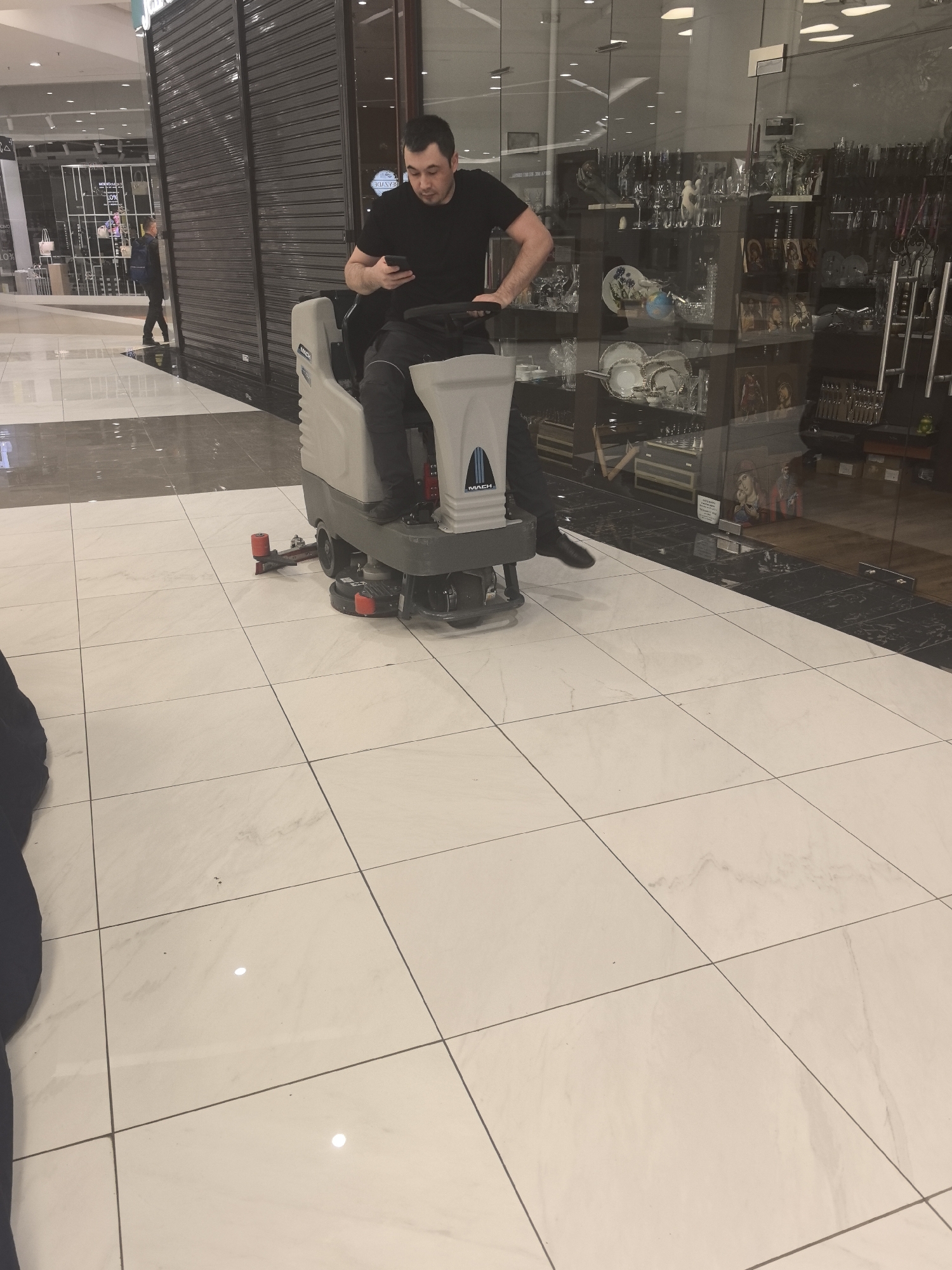 A man is sitting on a floor cleaning machine in a shopping mall. The man is wearing a black t-shirt and grey pants. He is holding a phone in his left hand. The floor cleaning machine is a light grey color. Behind the man and the machine is a store front. The store is closed with security doors. Other store fronts with glass are on the right. The floor is a white tile. Another person is walking in the far background. The location is a shopping mall in an unknown city.