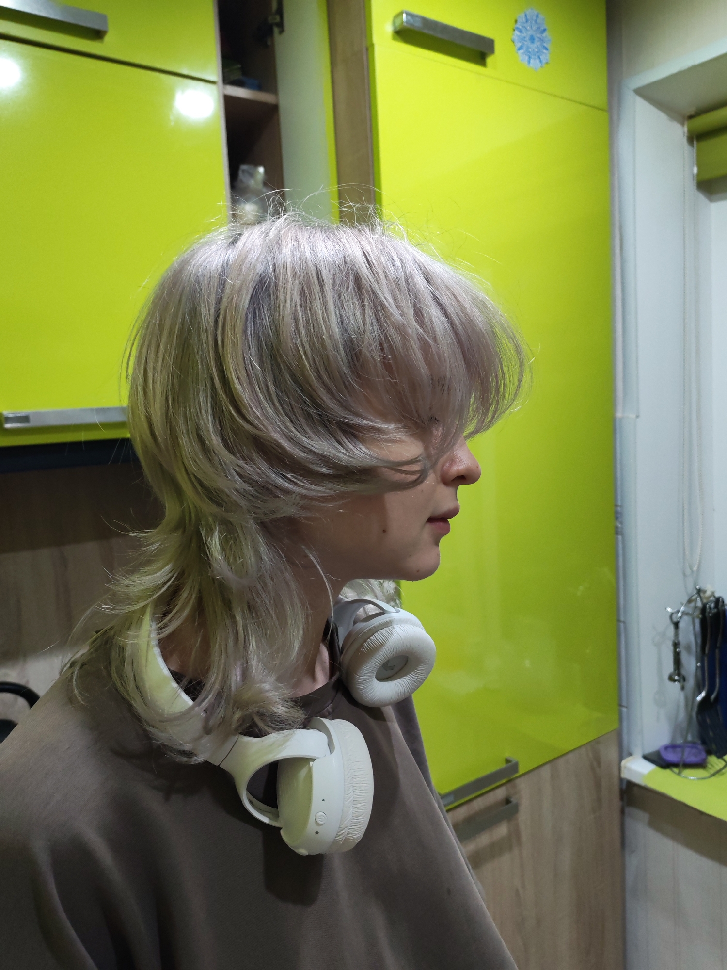 The image depicts a person with light-colored, layered hair and white headphones around their neck, positioned in profile. They are wearing a brown t-shirt. The background consists of bright lime-green cabinets with silver handles and a wooden cabinet. A small blue snowflake sticker is visible on the upper right cabinet. The scene appears to be indoors, possibly a kitchen or a room with custom cabinetry. The lighting suggests an indoor setting with artificial light sources. No specific location in Chaikovsky, Russia is identifiable from the image.