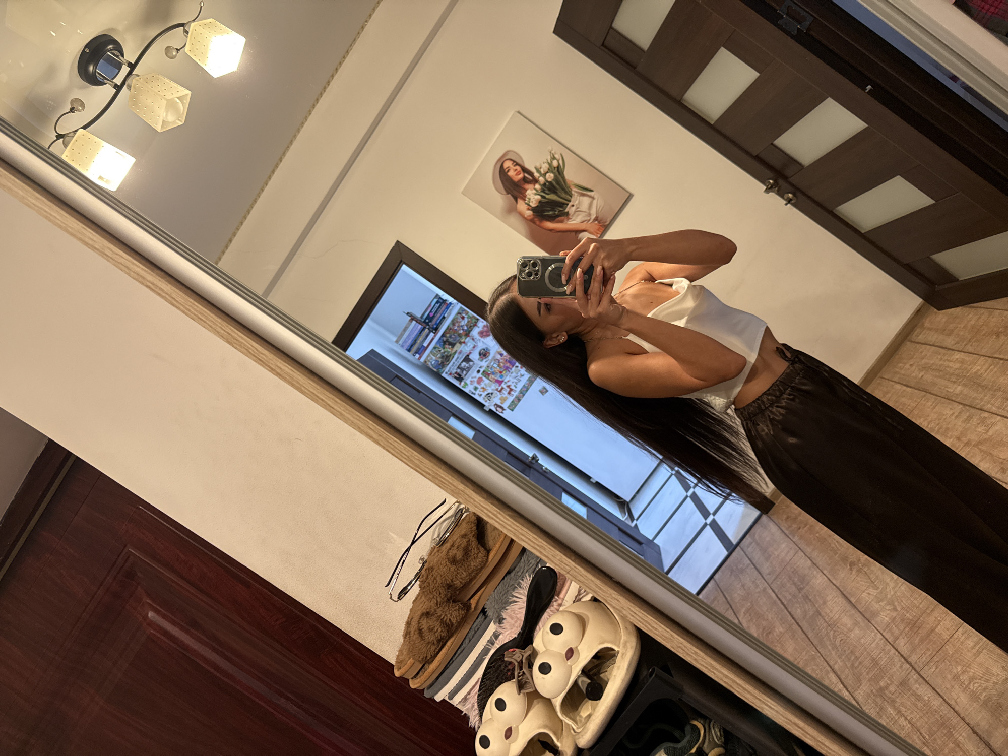 The image depicts a woman taking a mirror selfie in what appears to be a residential interior. She is wearing a white satin camisole and dark, loose-fitting pants. Her long, dark hair cascades down her back. In the foreground, reflected in the mirror, are a stack of items including slippers with cartoonish faces, a hairbrush, and a pair of glasses resting on folded fabric. The background shows a wooden door, a framed picture of a woman holding tulips, and a ceiling light fixture with three illuminated shades. The flooring is made of wood planks. There is no visible information in the image that would indicate a specific location within Chișinău, Moldova.