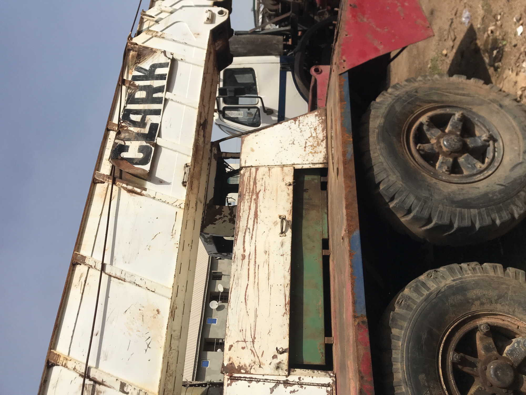 The image captures a heavily weathered, large truck, possibly a crane or specialized industrial vehicle, presented vertically against a clear, light blue sky. The truck's main body is white, heavily marked with rust streaks and patches on its metal panels and structural beams. Prominently displayed on one side panel are large, black, somewhat faded letters spelling "CLARK".
The truck's cabin, visible between parts of its structure, is also white with a blue stripe. Below the main body, two large, worn tires with dark, multi-spoke wheel hubs are visible, resting on a dusty, unpaved surface. One tire sidewall shows the text "TL 558" and what appear to be faint Chinese characters.
In the background, a multi-story building with several windows and satellite dishes can be seen, suggesting an urban or industrial environment. The scene depicts the truck parked and stationary during daylight hours. No people are visible, and no specific activity is occurring. The extensive rust and wear indicate significant age and heavy use of the vehicle.
