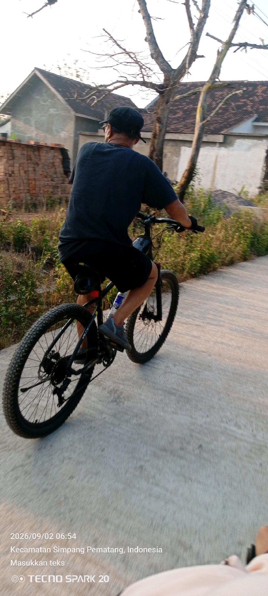 A person is riding a bicycle on a paved road in Simpang Pematang, Indonesia. The person is wearing a dark t-shirt, black shorts, a black cap, and grey shoes. The bicycle is black and has knobby tires. In the background, there are houses, trees, and vegetation. The image is timestamped 2026/09/02 06:54. The text "Kecamatan Simpang Pematang, Indonesia" and "Masukkan teks" and "TECNO SPARK 20" are overlaid on the bottom of the image.