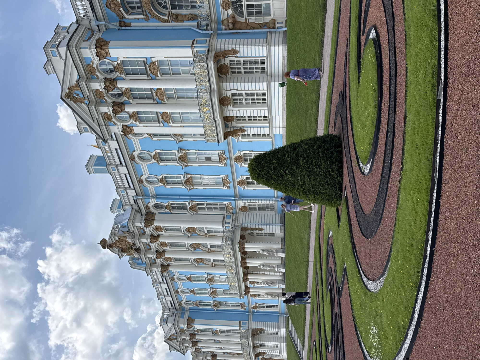 In Pushkin, Russia, the Catherine Palace, a grand structure painted in bright blue with white and gold architectural details, stands tall. In front of the palace, a formal garden features intricate swirling patterns of grass and dark-colored ground cover, bordered by a reddish-brown mulch. A conical evergreen tree is positioned in the foreground. Several people are present, with two individuals walking near the palace entrance on the left side of the image. Further to the right, a woman in a blue and white patterned dress walks along a path, holding a red object, while a man stands nearby on the lawn. The sky above is a mix of blue and scattered white clouds.