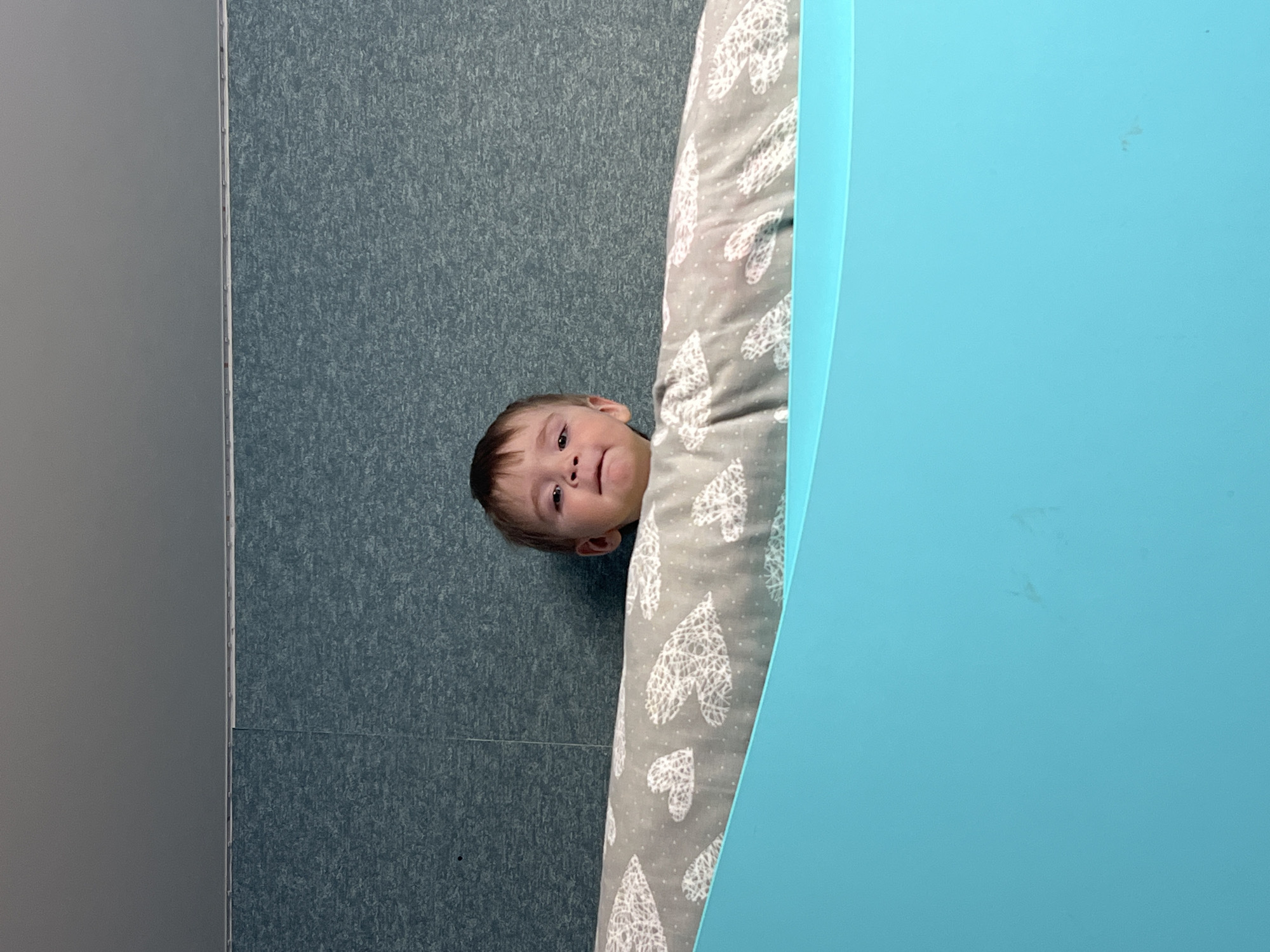 A young child is partially visible behind a patterned cushion and a light blue table. The child is looking at the camera. The background consists of a grey wall. The exact location within the city of Noviy, Russia is not discernible from the image.