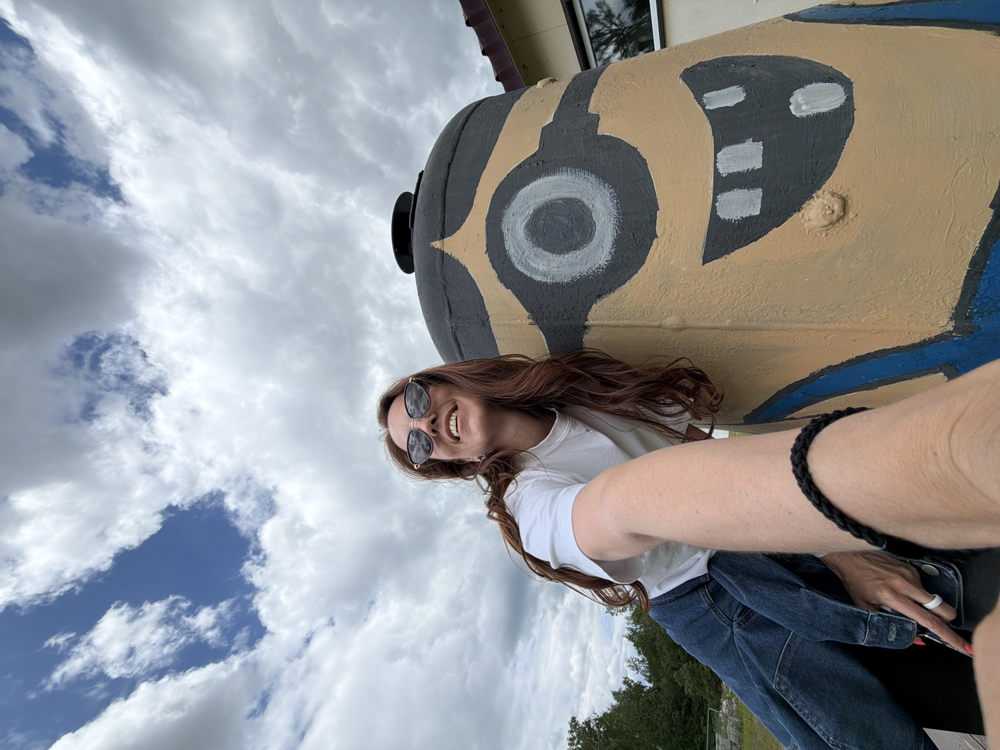 A woman with long, auburn hair and sunglasses is taking a selfie outdoors. She is wearing a white t-shirt and denim jacket, and leaning against a large, cylindrical object painted to resemble a Minion character. The object has a single large eye and a wide, smiling mouth. The background consists of a cloudy sky and some trees. This photo appears to be taken in Birobidzhan, Russia.