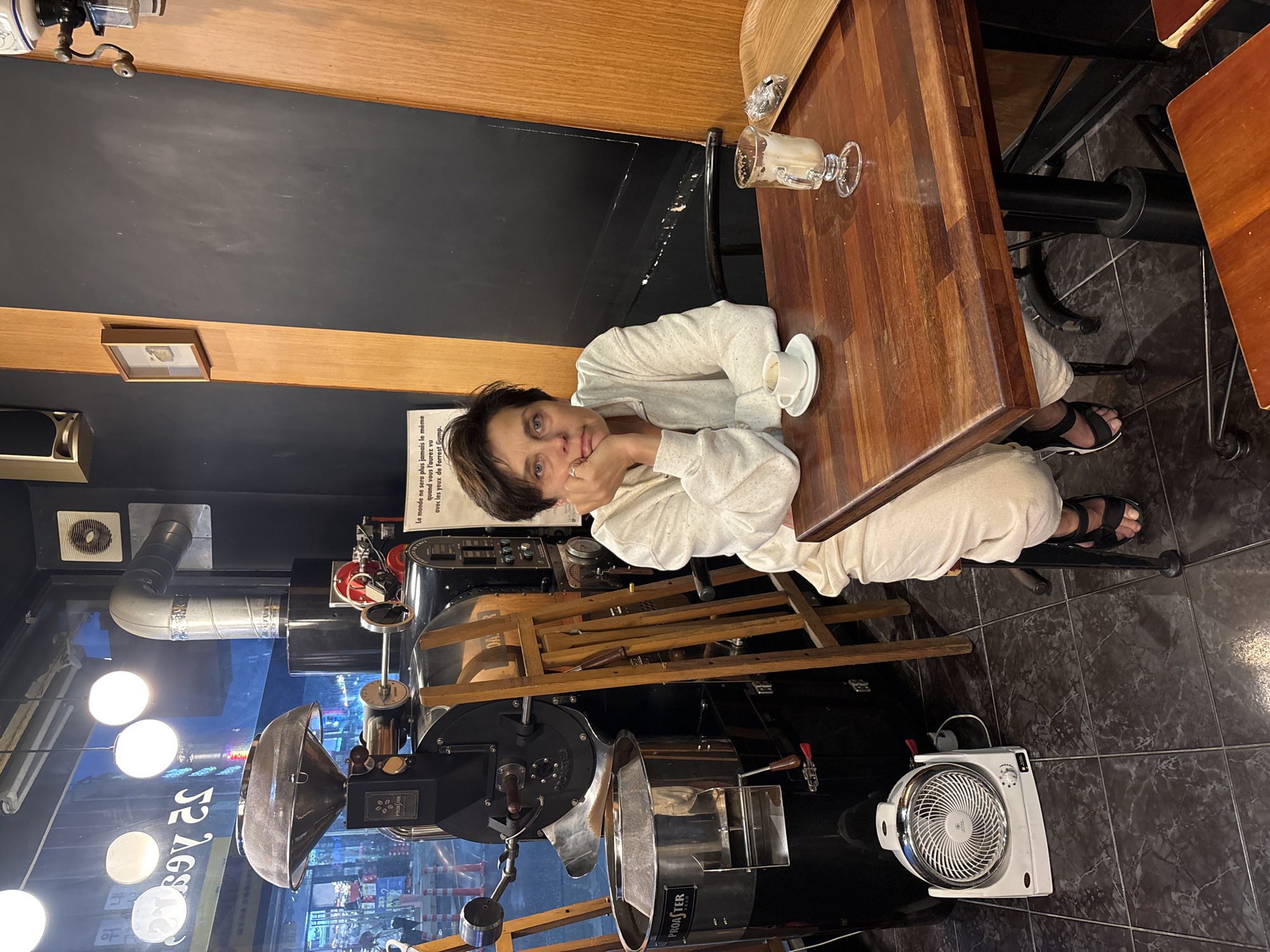 A person is sitting at a wooden table in a dimly lit establishment, possibly a coffee shop or roastery, based on the presence of a large coffee roasting machine in the background. The person is leaning on the table, resting their head on their hands, with a pensive expression. They are wearing a light-colored sweater and pants. On the table are a cup and saucer, and a glass with a drink, suggesting they are enjoying a beverage. The overall ambiance appears relaxed and intimate. A sign in French, with the text "Le monde ne sera plus jamais le même quand vous l'aurez vu avec les yeux de Forrest Gump," is visible on the wall behind the person, which translates to "The world will never be the same again when you have seen it through the eyes of Forrest Gump." The coffee roasting machine is a prominent feature, indicating a focus on coffee production. Other objects like lights and a fan are also visible, adding to the industrial yet cozy atmosphere of the place. The image seems to be taken indoors, likely during the evening or at a time when the lighting is subdued.