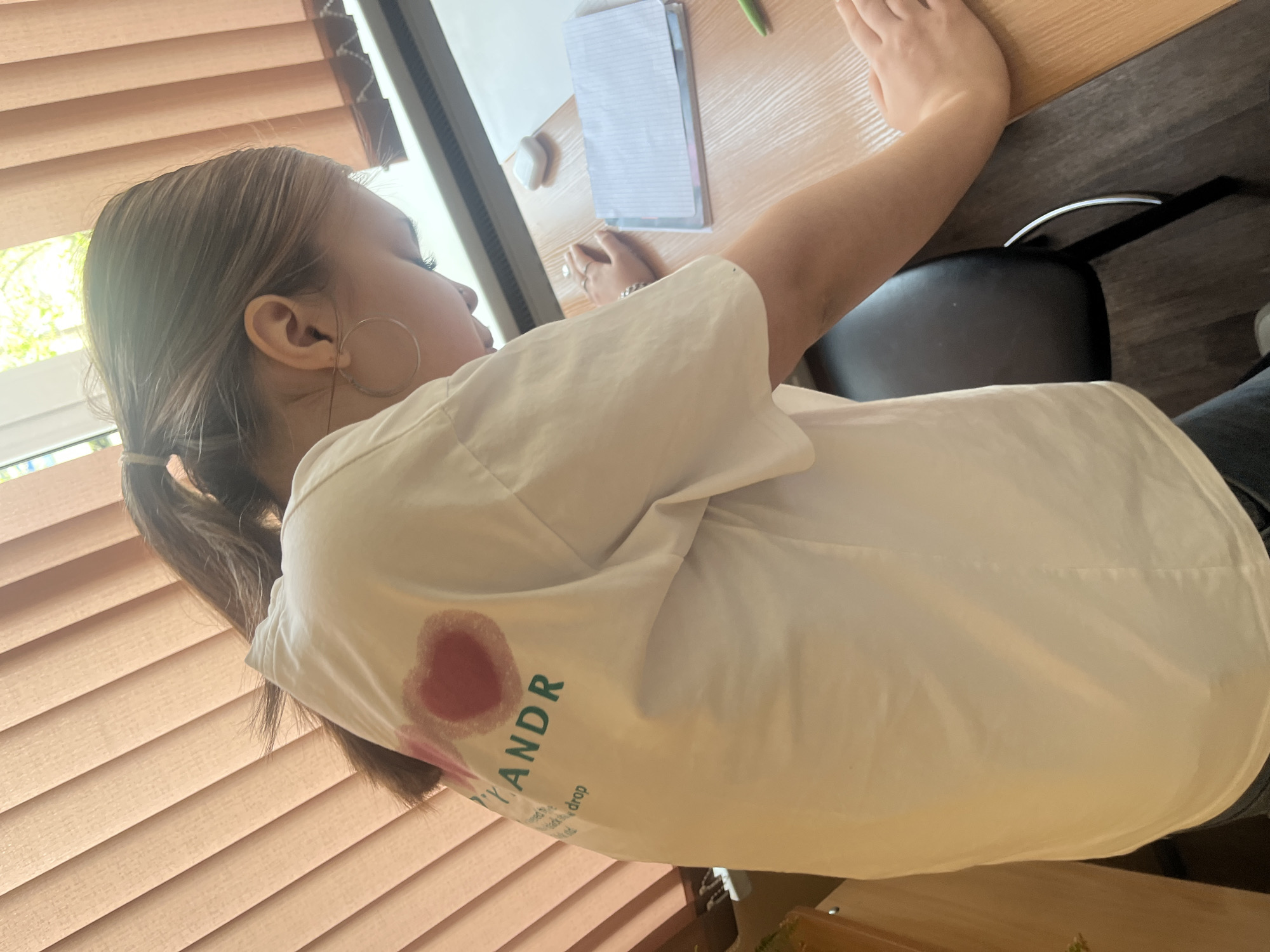 A woman with brown hair in a low ponytail and wearing hoop earrings is seated at a desk. She is wearing a white t-shirt with text and a heart-shaped graphic in pink and red on the back. She is writing on a pad of lined paper on the desk. A green pen is also on the desk. A black chair is behind her. The desk is light brown with a dark brown edge, and is set in front of a window covered by closed, horizontal blinds. The location cannot be confirmed as Saint Petersburg, Russia.